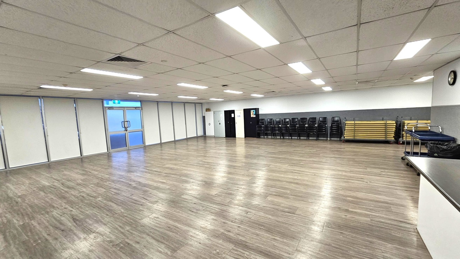 A spacious room with a wooden floor and a grid-patterned ceiling with fluorescent lights. On the left, there are large windows with blinds and a double glass door. The back wall has a stack of black chairs and folded tables. On the right, a clock is mounted on the wall above a counter with a trolley and a bin. The room appears to be a multipurpose space, possibly for meetings or events.