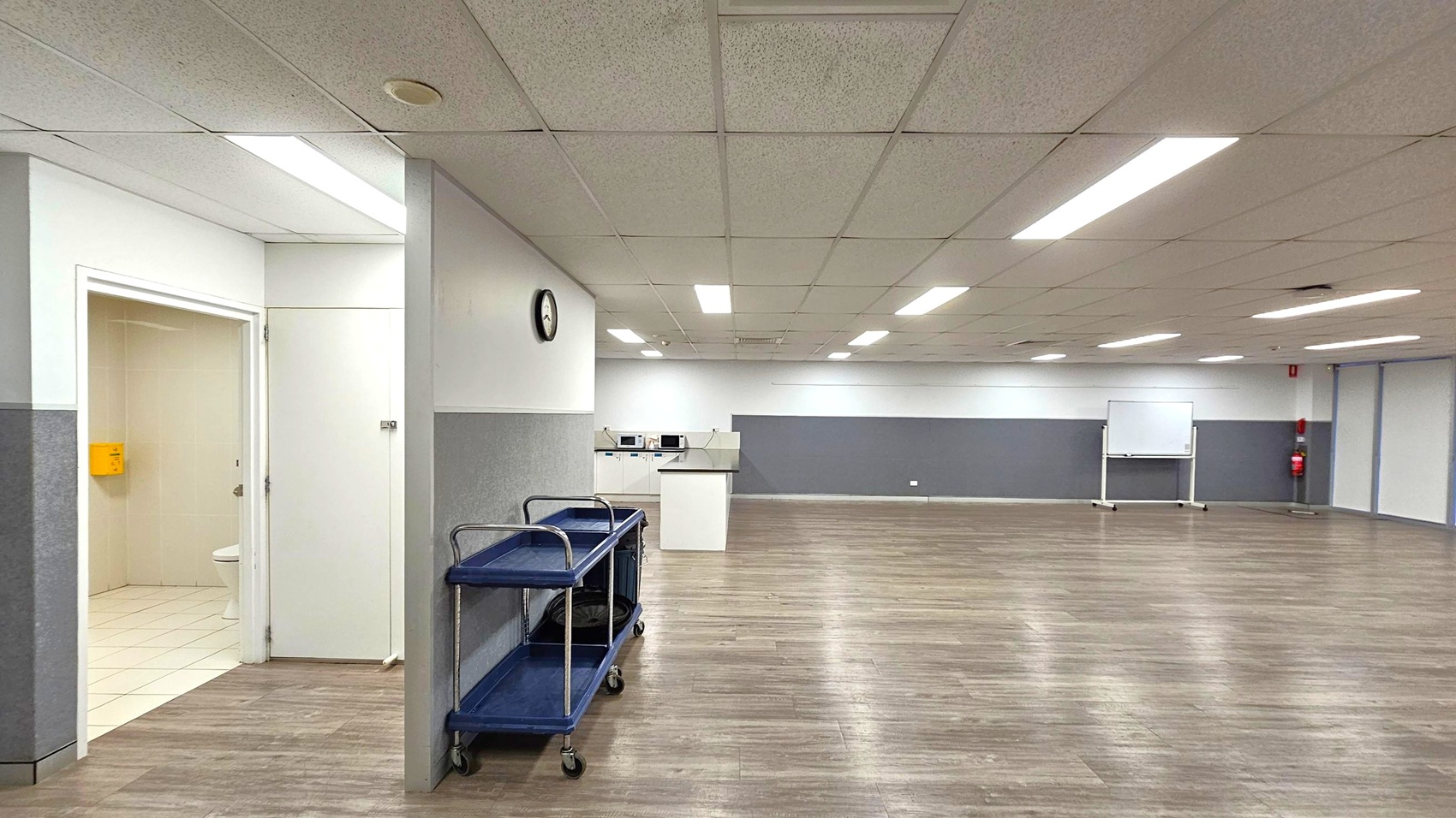 A spacious room with a light-coloured tiled floor and a suspended ceiling with fluorescent lights. On the left, there's an open door leading to a small room with a toilet. A dark trolley with shelves is positioned against the wall, which has a clock mounted on it. In the background, there are several microwaves on a counter and a whiteboard on wheels. The walls are grey and white, and the room has a clean, functional appearance.