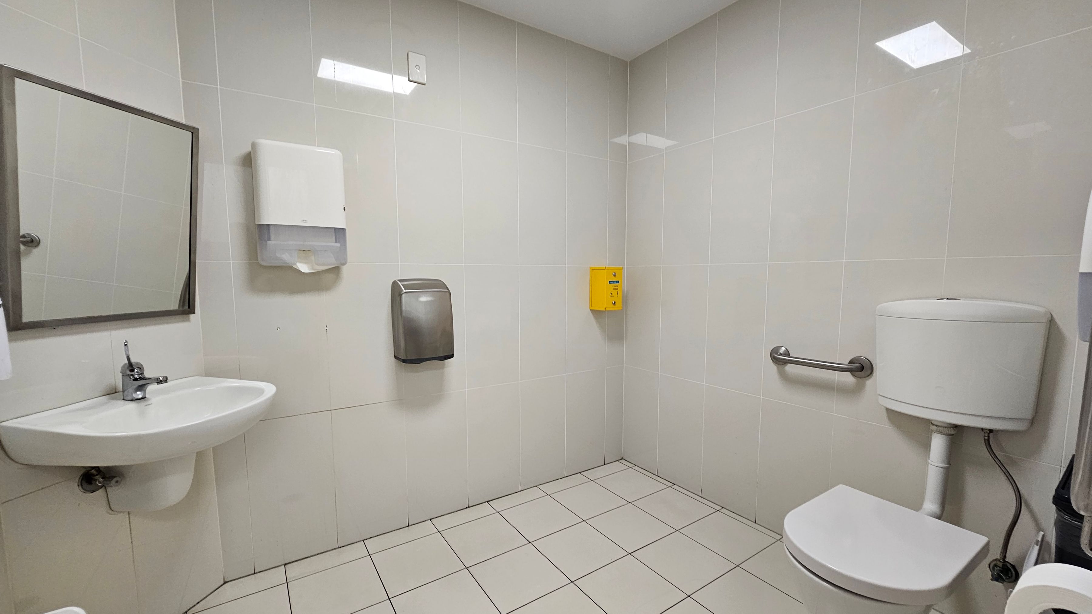 A clean, white-tiled bathroom with a sink and mirror on the left. Above the sink is a paper towel dispenser, and next to it, a hand dryer. On the right, there's a toilet with a grab bar beside it. A small yellow box is mounted on the wall above the toilet. The floor is tiled in white, and the ceiling has recessed lighting.