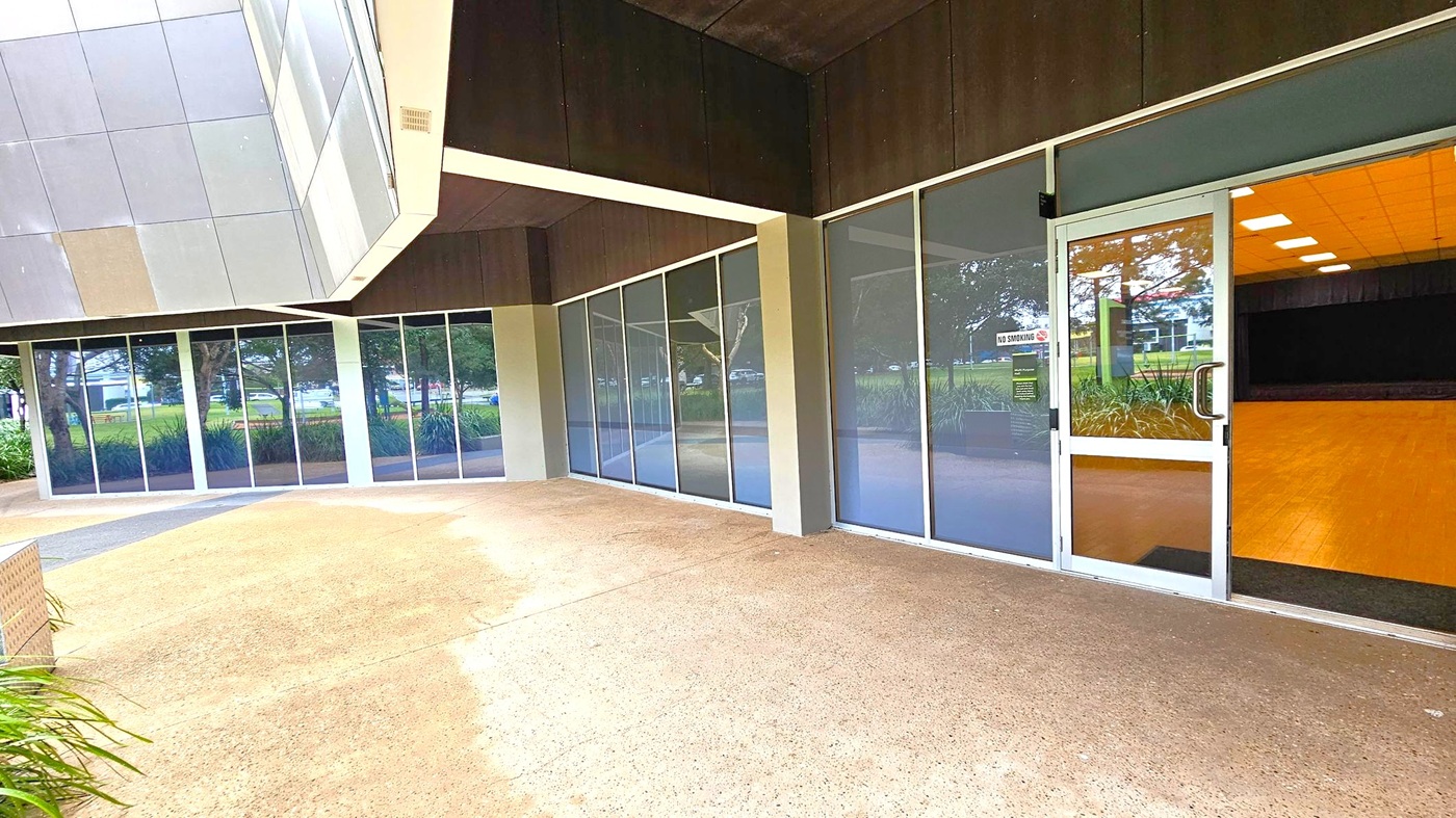 A modern building exterior with large glass windows and doors. On the right, a glass door reveals an interior with a wooden floor and ceiling lights. The left side features reflective glass panels showing trees and greenery outside. The ground is a textured concrete surface, and part of the building's geometric facade is visible above.