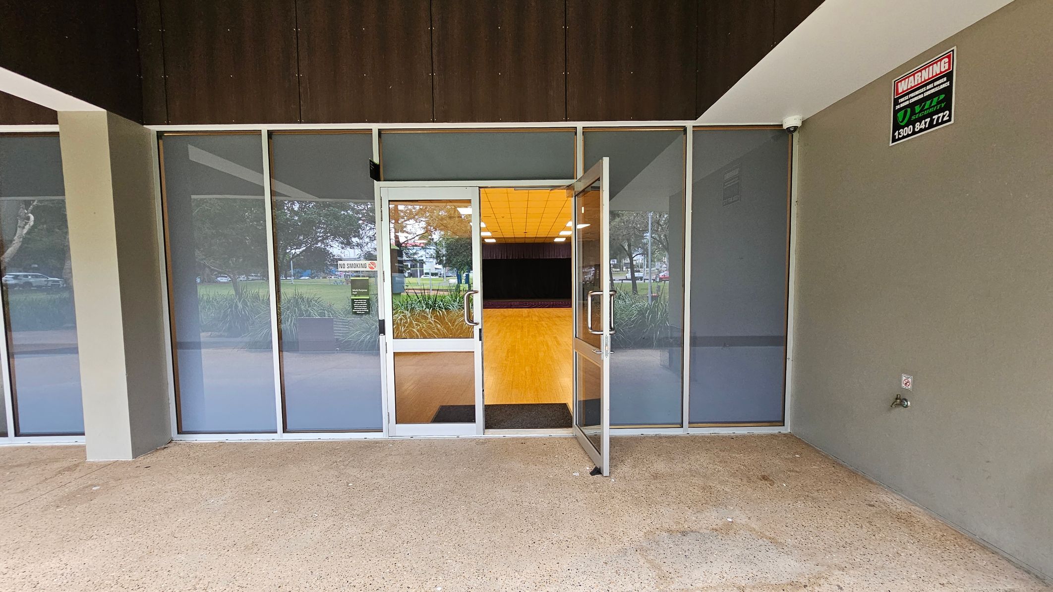 A glass entrance with double doors, one of which is open, revealing an interior with a wooden floor and a ceiling with lights. The exterior has a textured concrete floor and grey walls. A warning sign is mounted on the right wall. Through the glass, there are visible trees and a grassy area outside.