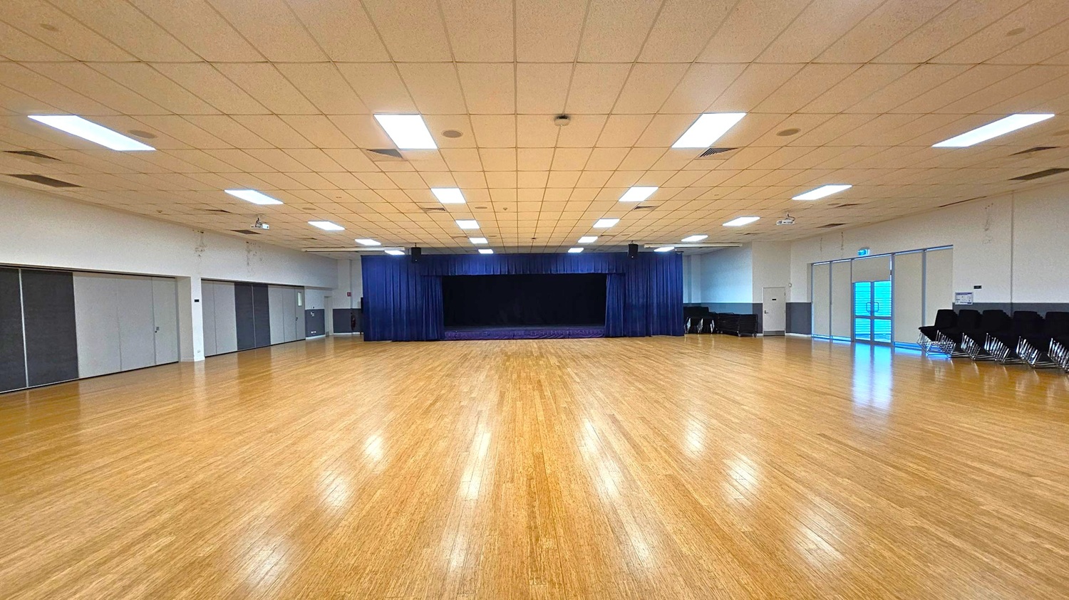 A large, empty hall with a polished wooden floor. In the centre, a stage with dark blue curtains is visible. The ceiling is lined with rectangular fluorescent lights. On the left, there are several closed, grey partition doors. On the right, stacked chairs are placed against the wall near a set of glass doors. The walls are plain and light-coloured, contributing to the spacious feel of the room.