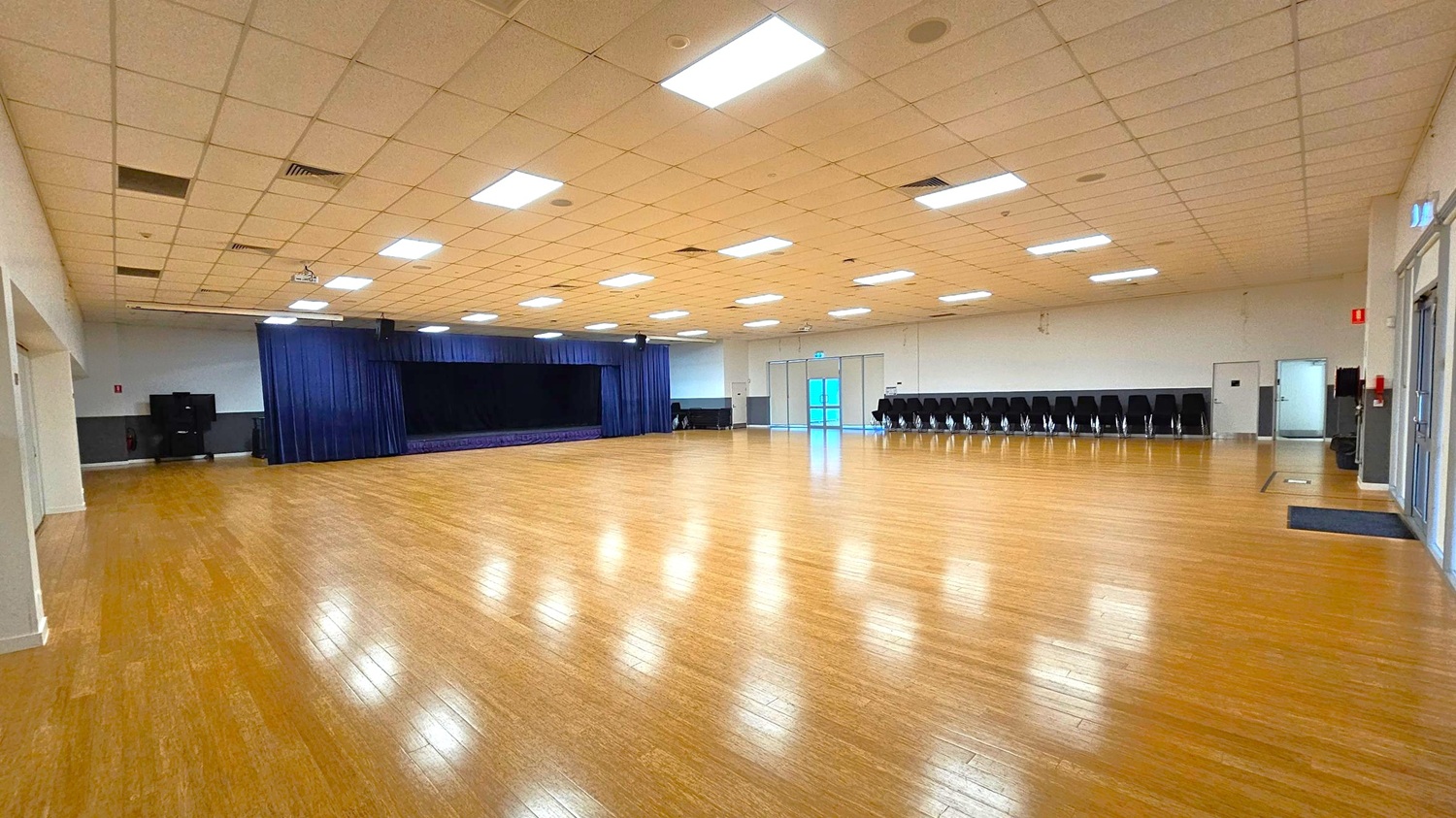 A large, empty hall with a polished wooden floor and a grid-patterned ceiling with fluorescent lights. On the left, a stage with dark blue curtains is visible. The right side features a row of stacked chairs against the wall. At the far end, there are double doors with glass panels. The walls are light-coloured, and there are a few doors and a fire extinguisher on the right.