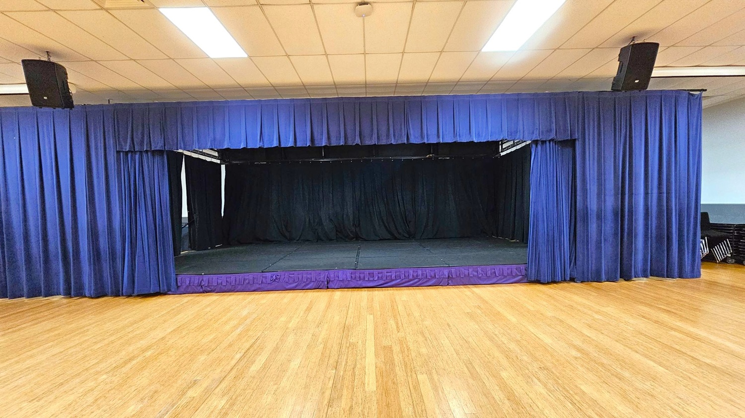 A stage with closed blue curtains is centred in the image. The stage floor is black, and the surrounding area has a light wooden floor. The ceiling is white with rectangular fluorescent lights. On the right, there are stacked chairs against the wall. Two black speakers are mounted at the top corners of the stage.