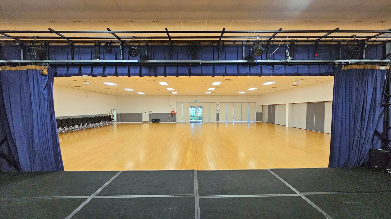 View from a stage with black flooring, looking out into a large hall. The stage is framed by blue curtains and overhead lighting equipment. The hall has a light wooden floor and a ceiling with recessed lighting. On the left, a row of stacked black chairs lines the wall. At the far end, there are glass doors with a view outside, flanked by a fire extinguisher and a clock. The right wall features large, grey sliding panels.