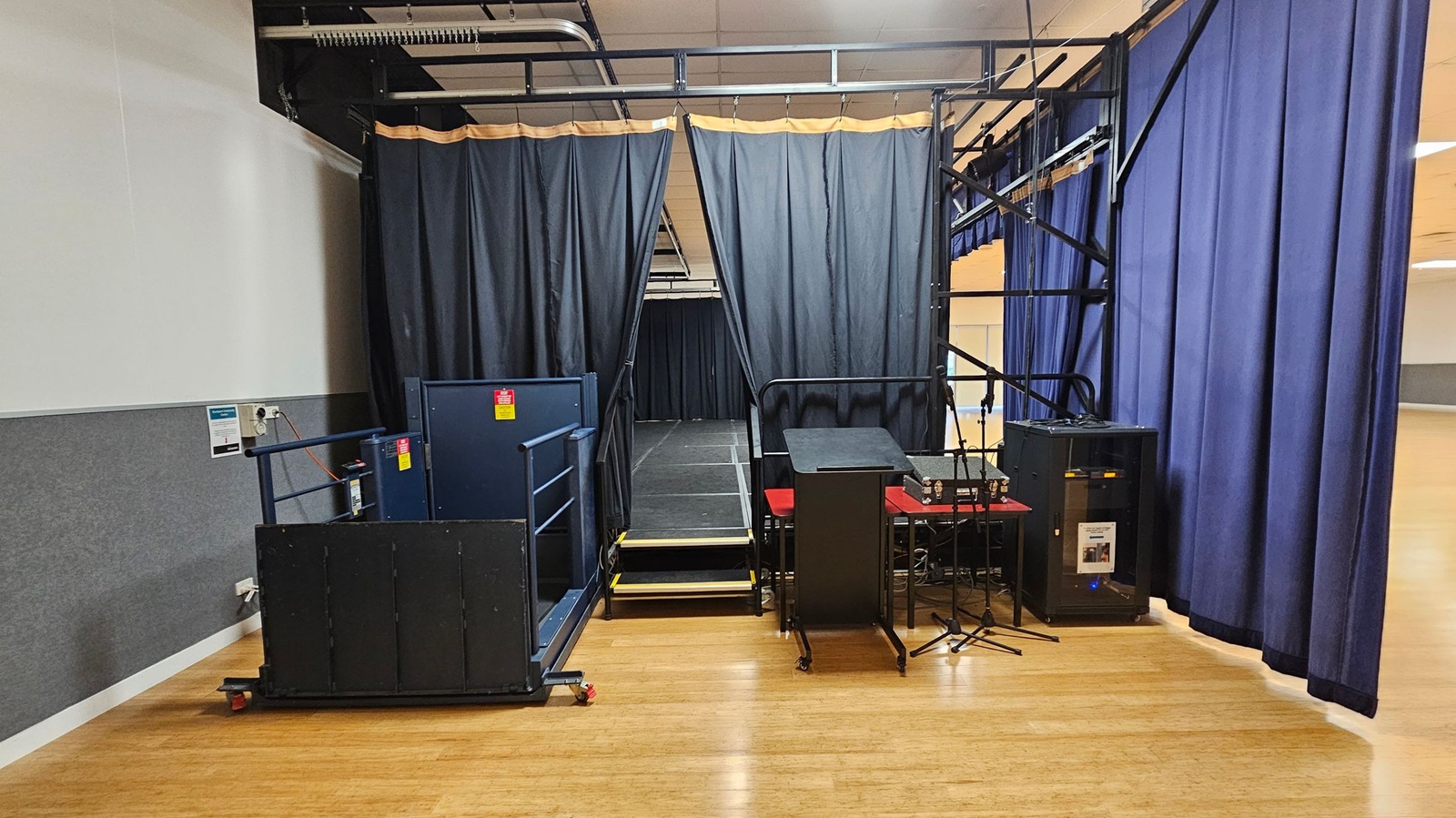 A small stage setup with black curtains in the centre, partially open to reveal a platform. On the left, a blue lift with railings is positioned near the stage. On the right, a podium on wheels, a microphone stand, and audio equipment are visible. The floor is wooden, and the ceiling has visible pipes and lighting fixtures. Blue curtains hang on the right side, partially covering the wall.