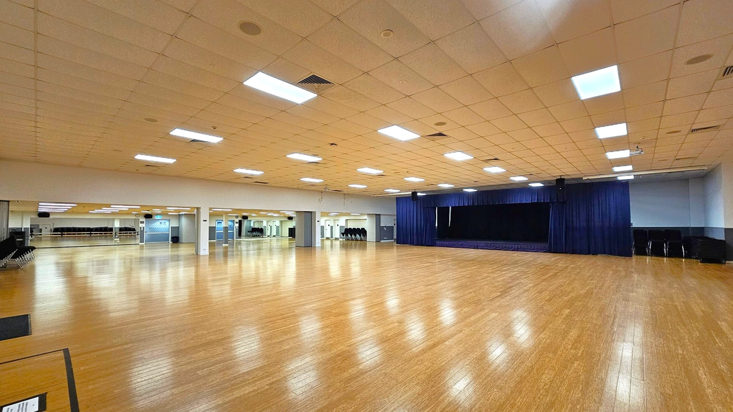 A large, empty hall with a polished wooden floor. On the right, a stage with dark curtains. The left side features a wall of mirrors, reflecting the room's interior. The ceiling is lined with rectangular fluorescent lights, and there are several columns throughout the space. Stacks of chairs are visible against the walls.