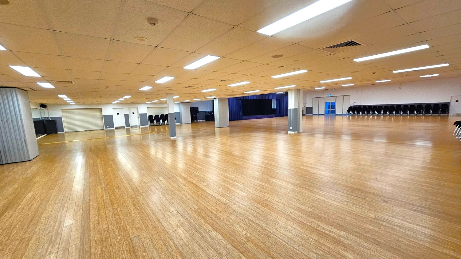A large, empty hall with a polished wooden floor and a grid-patterned ceiling with fluorescent lights. In the background, there is a stage with a dark curtain. On the left, a closed roller shutter and a stack of chairs are visible. On the right, a row of stacked chairs lines the wall, and a set of double doors with blue frames is present. Several support columns are evenly spaced throughout the room.