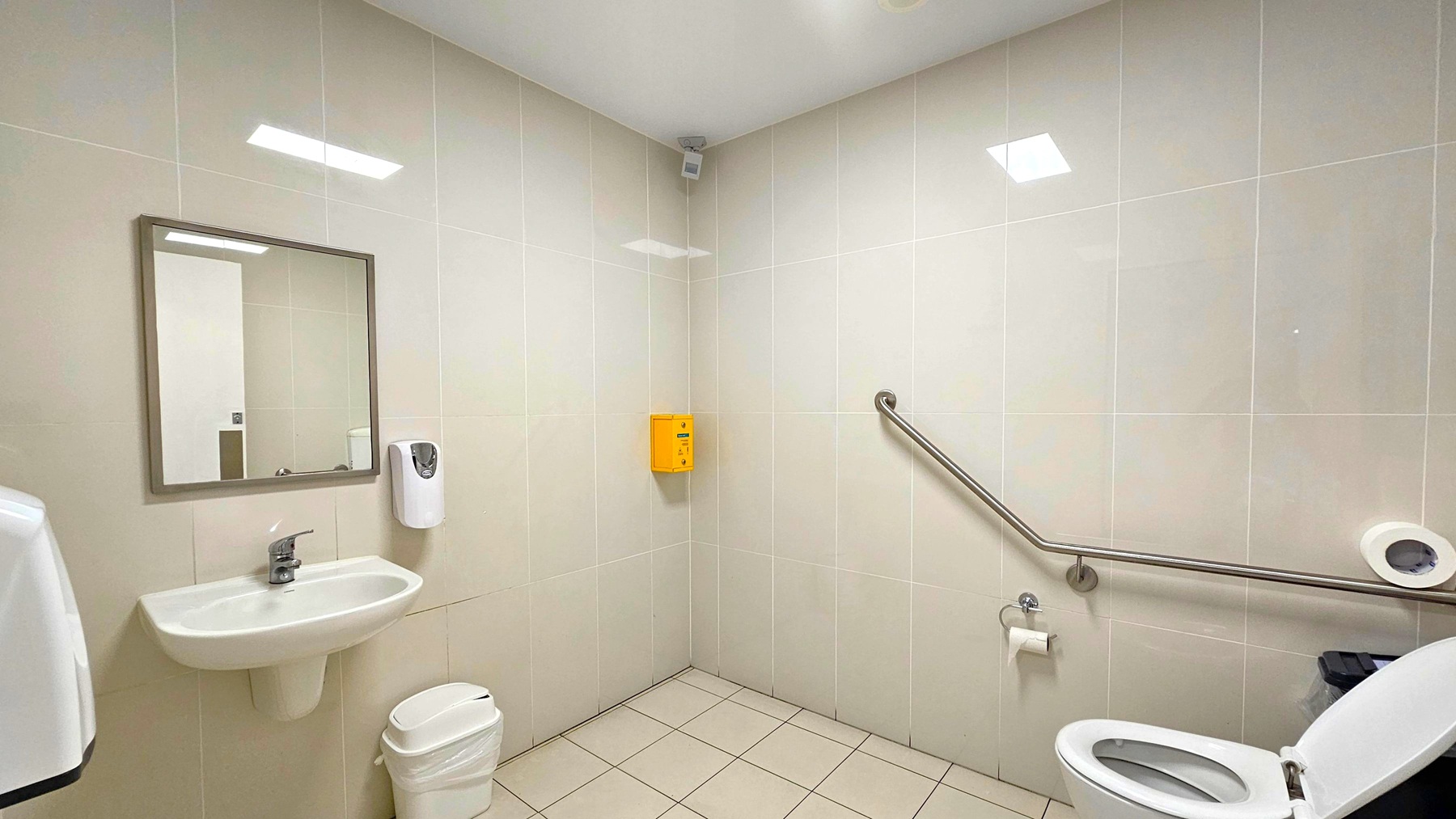 A clean, accessible bathroom with white tiled walls and a light-coloured tiled floor. On the left, a wall-mounted sink with a mirror above and a soap dispenser to the right. Below the sink is a small waste bin. On the right, a toilet with an open lid and a grab rail along the wall. A toilet paper holder is mounted nearby. A yellow box is fixed to the wall in the corner. The ceiling is plain white with recessed lighting.