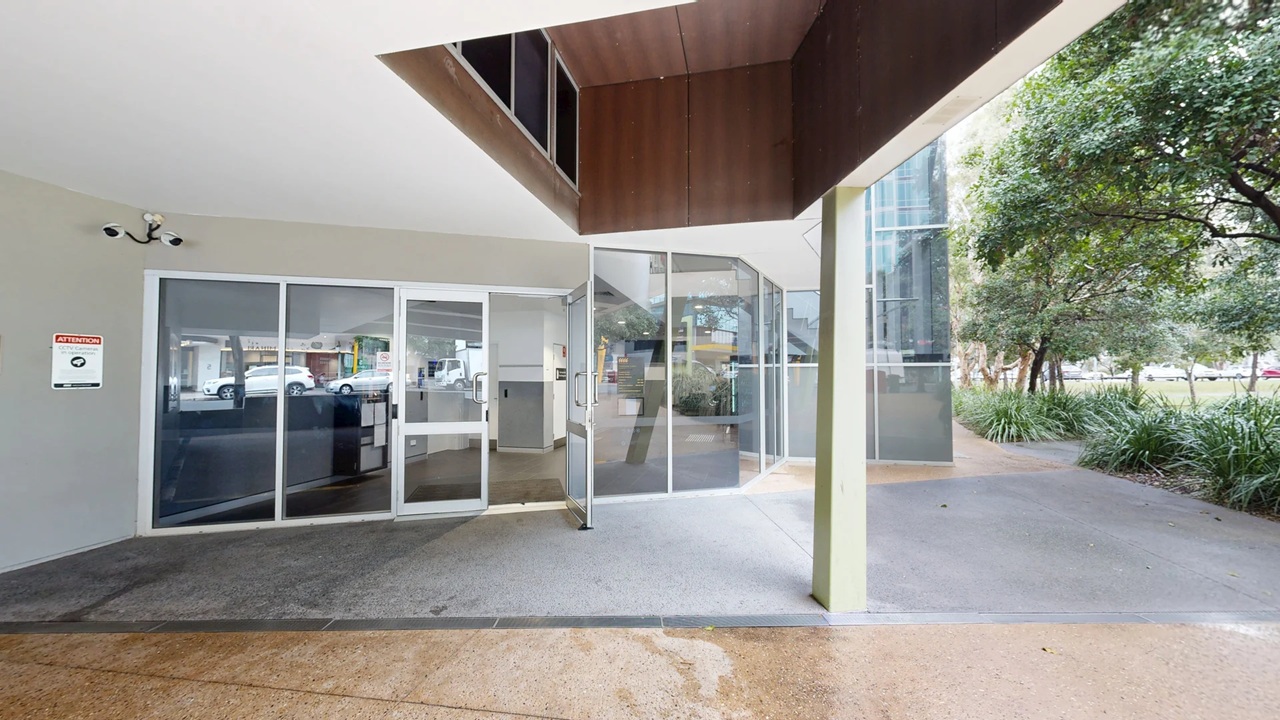 A modern building entrance with large glass doors and windows on the left. The floor is a mix of concrete and tiled surfaces. On the right, there is a pathway leading to a garden area with trees and shrubs. The ceiling is white with a wooden panel section above the entrance. A security camera is mounted on the wall near a sign.