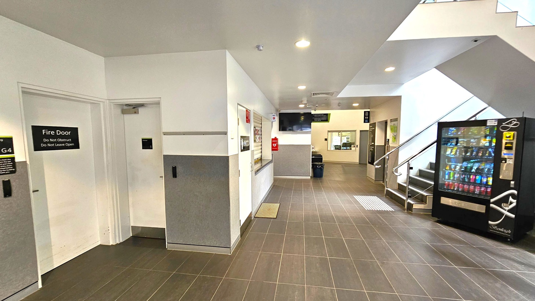 A hallway with brown tiled flooring and a white ceiling. On the left, there is a fire door with a sign, and a service window with a closed shutter. In the centre, an AED is mounted on the wall. On the right, a vending machine is next to a staircase with metal railings leading upwards. Further back, there is a reception area with a window and a counter. The space is well-lit with ceiling lights.