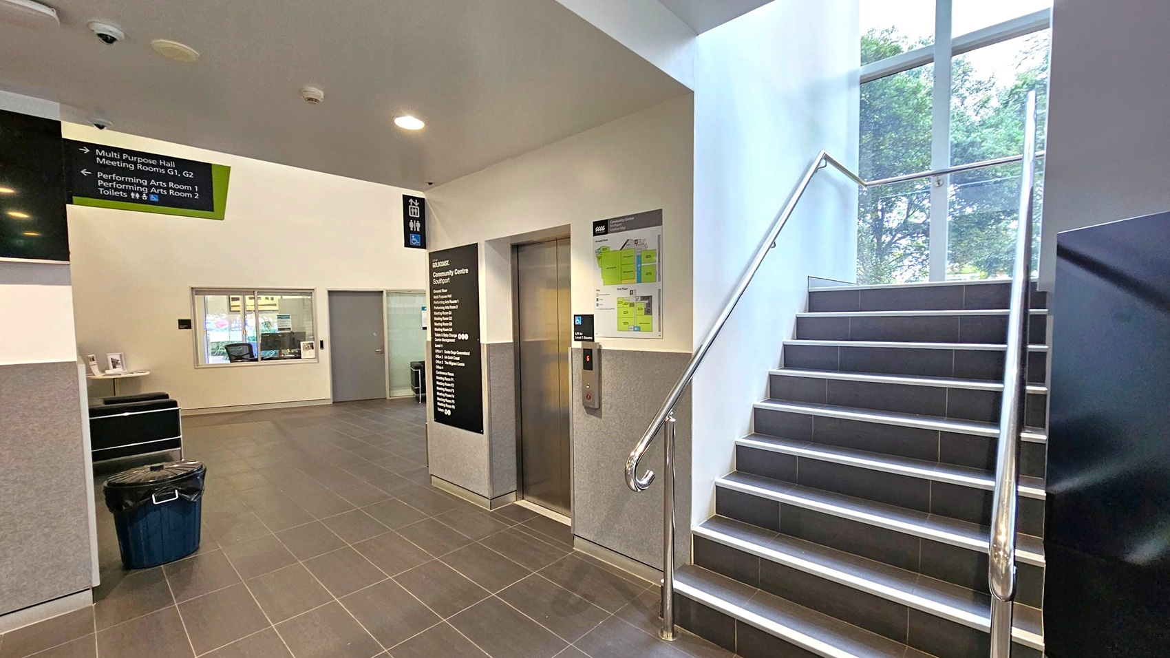 A modern indoor space with a tiled floor and a white ceiling featuring recessed lighting. On the right, a staircase with metal railings leads upwards, adjacent to large windows showing trees outside. In the centre, there's a lift with a control panel and a directory board. On the left, a wall-mounted sign provides directions to various rooms. A small reception area with a window and a door is visible, along with a bin placed on the floor.
