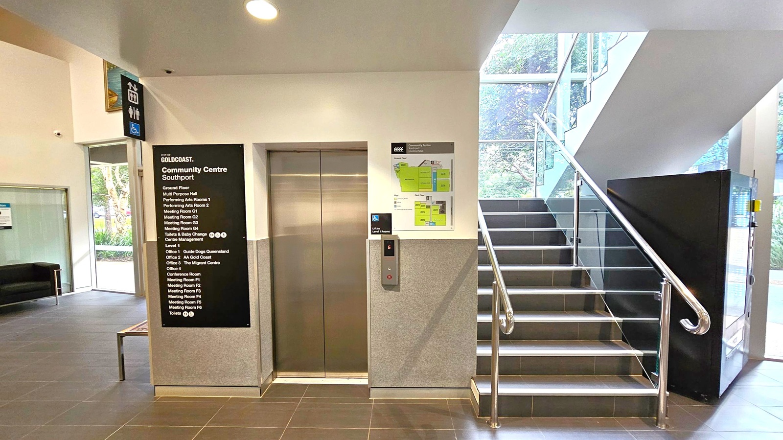Elevator doors are in the centre with a directory sign on the left listing various rooms and offices in the community centre. A staircase with metal railings is on the right, leading upwards. A vending machine is positioned next to the stairs. The floor is tiled in dark grey, and the ceiling is white with recessed lighting. Large windows in the background allow natural light to enter, revealing greenery outside.