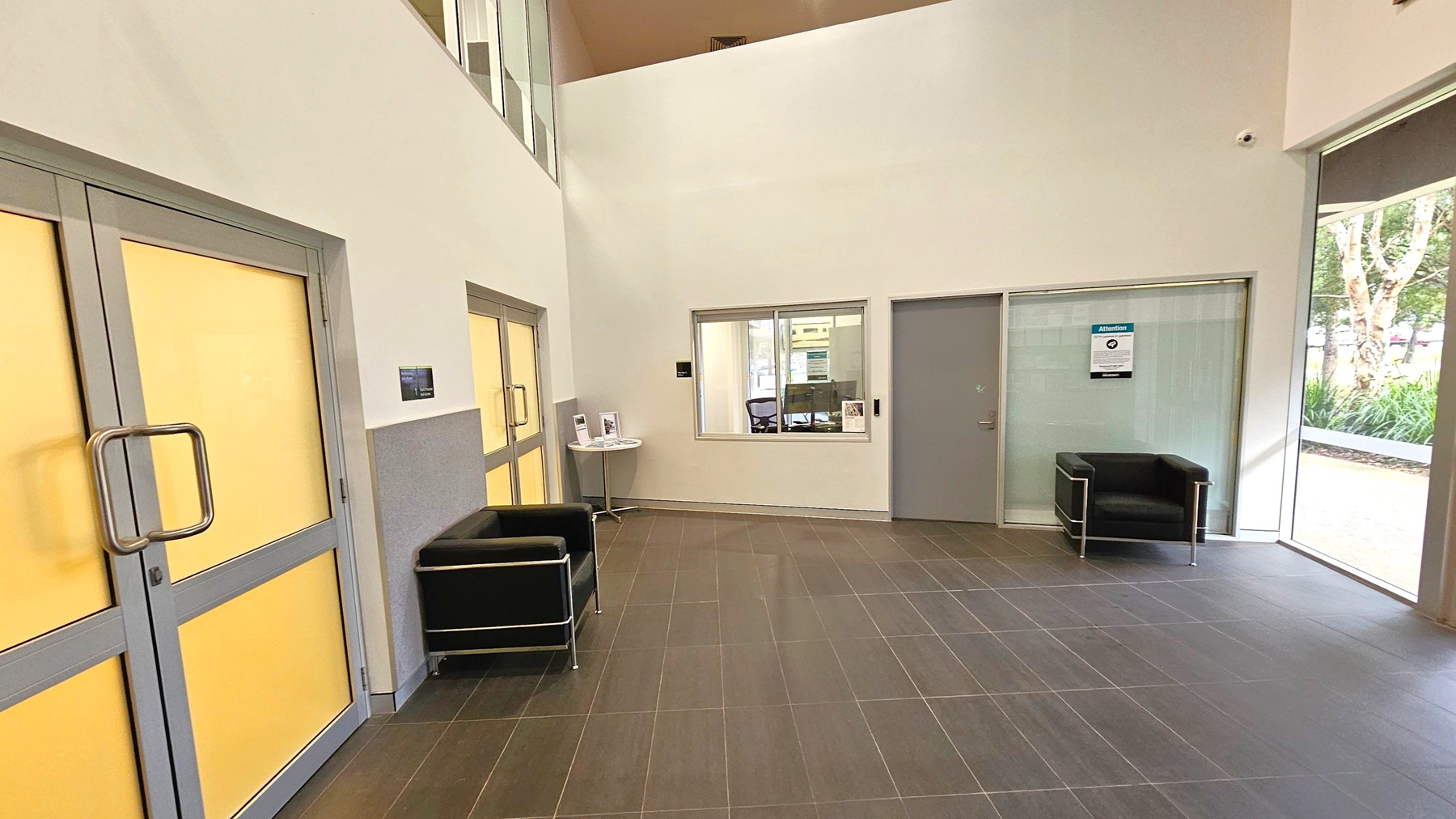 A spacious interior with a high ceiling and tiled floor. On the left, there are double doors with yellow panels and a black armchair. In the centre, a small table with brochures is placed near a window that looks into another room. To the right, a grey door and another black armchair are positioned near large windows showing greenery outside.