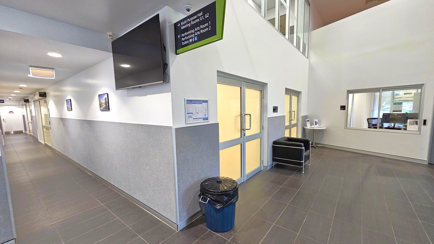 A hallway with grey tiled flooring and white walls. On the left, a corridor extends with several doors and framed pictures on the wall. A large black screen is mounted above. In the centre, double doors with frosted glass are flanked by a wall-mounted sign and a black bin. On the right, a window looks into an office space with a chair and a small table displaying brochures. The ceiling is white with recessed lighting.