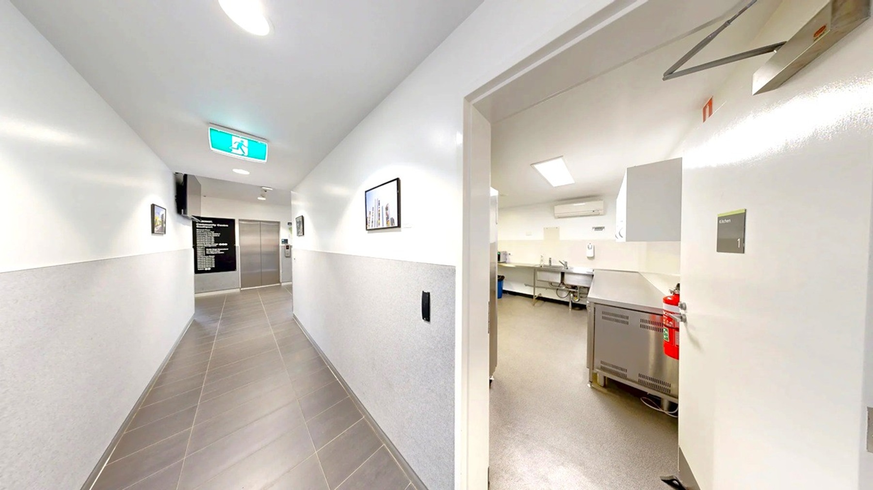 A corridor with white walls and a grey tiled floor. On the left, there are framed pictures on the wall and an illuminated exit sign on the ceiling. At the end of the corridor, a black directory board is visible next to a lift. On the right, an open door leads to a room with white walls, a beige floor, and visible kitchen equipment, including a stainless steel counter and a fire extinguisher. The ceiling has recessed lighting.