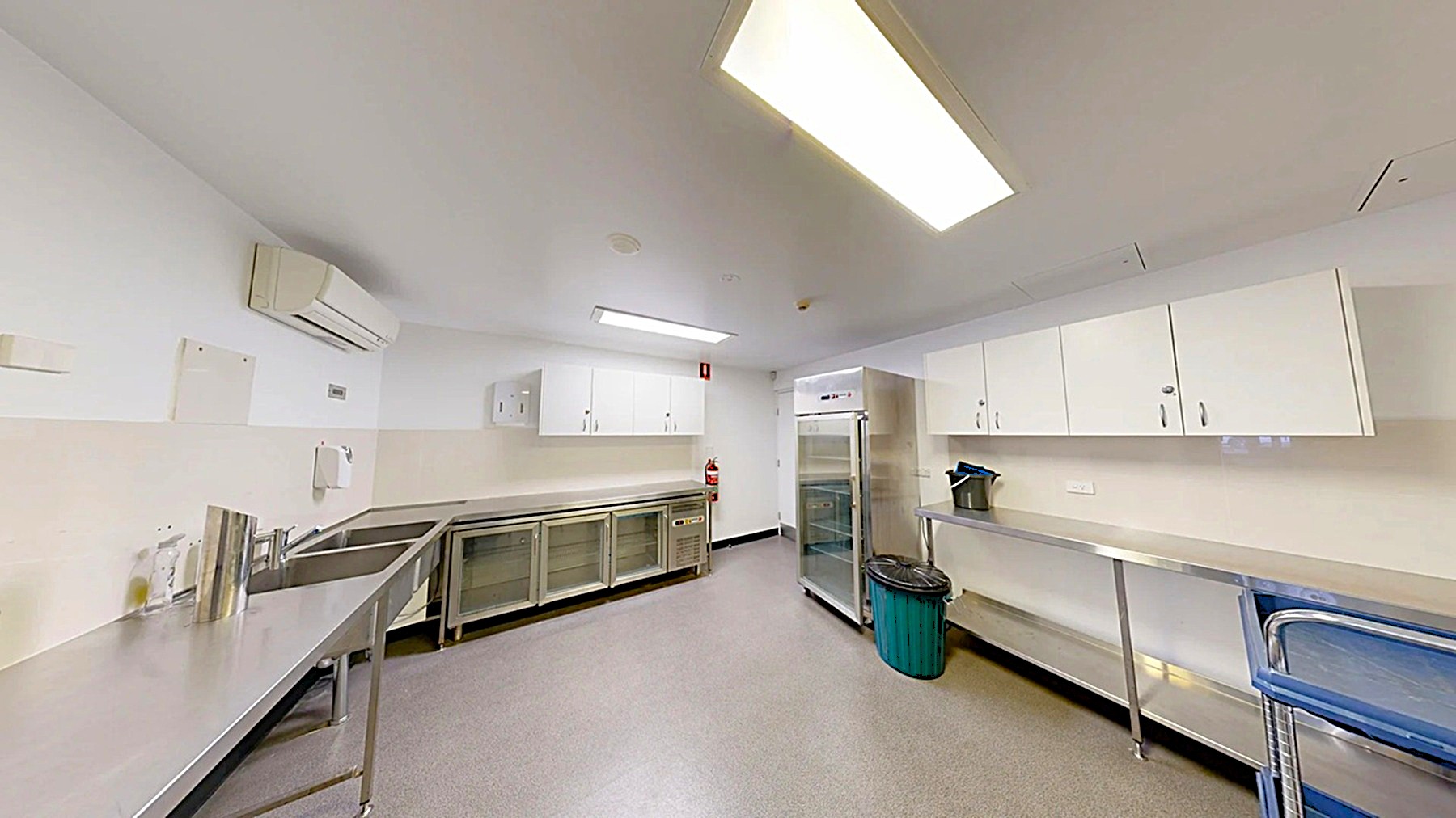 A commercial kitchen with stainless steel worktops on both sides. On the left, there are metal shelves and an air conditioning unit above. The right side features white cabinets above the worktop, a green bin, and a trolley. In the centre, a large stainless steel refrigerator stands next to a fire extinguisher. The floor is grey, and the ceiling has fluorescent lighting.