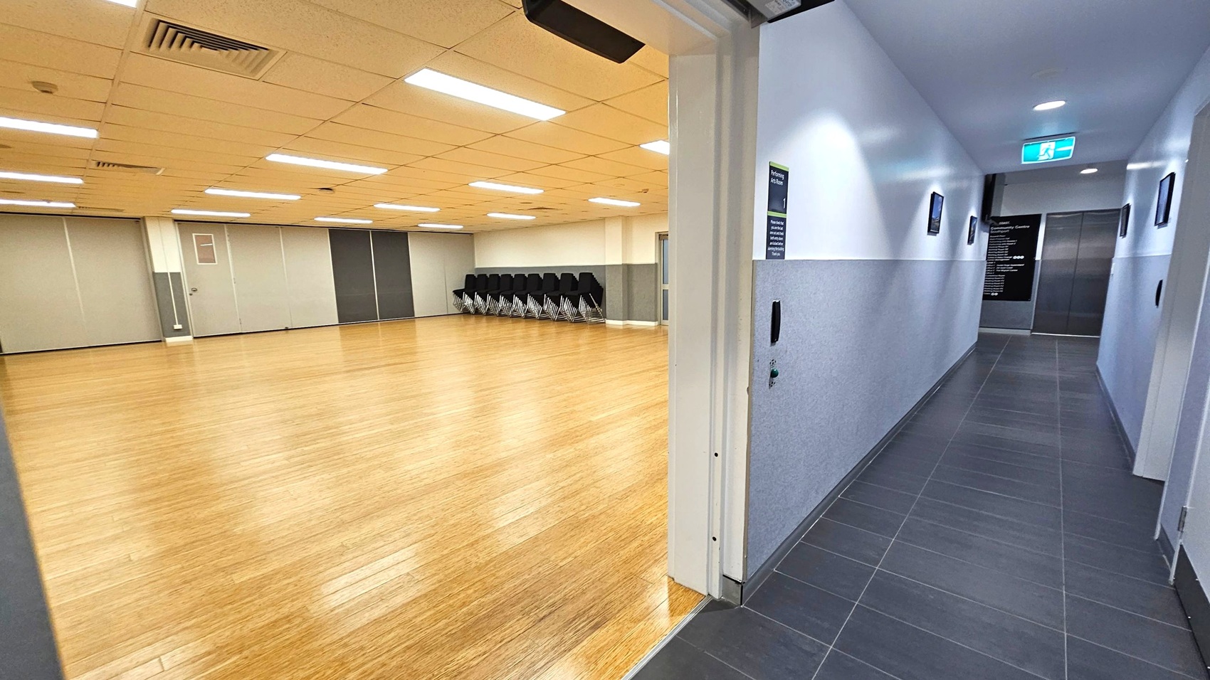 On the left, a spacious room with a wooden floor and a ceiling with fluorescent lights. Stacked chairs are against the far wall. On the right, a corridor with dark tiled flooring and white walls, leading to an elevator. An exit sign is above the corridor, and there are small signs on the walls. The ceiling in the corridor has recessed lighting.
