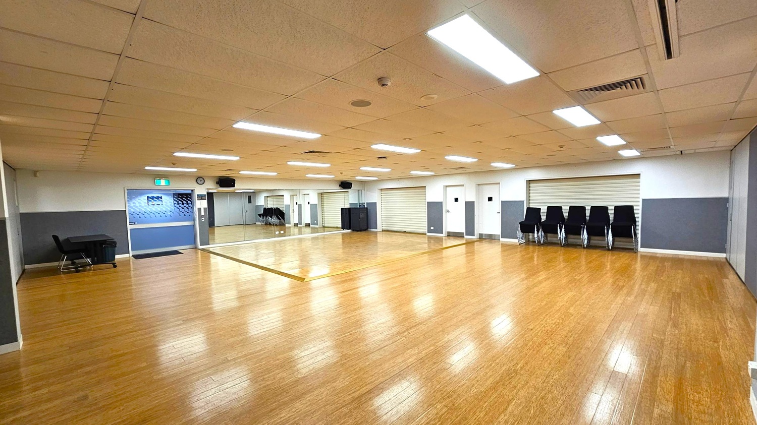A spacious room with a wooden floor and a ceiling with fluorescent lights. On the left, there is a black table and chair near a blue door with an exit sign above it. The back wall features large mirrors and a clock. On the right, several stacked black chairs are placed against a wall with a closed roller shutter. The walls are painted in grey and white, and the room appears to be well-lit and organised.