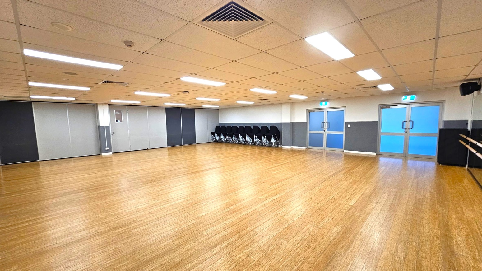 A spacious room with a wooden floor and a ceiling with fluorescent lights. On the left, there are grey and black partition panels. In the middle, a stack of black chairs is neatly arranged against the wall. On the right, there are two sets of double doors with blue-tinted glass and exit signs above them. A mirrored wall with ballet barres runs along the right side.