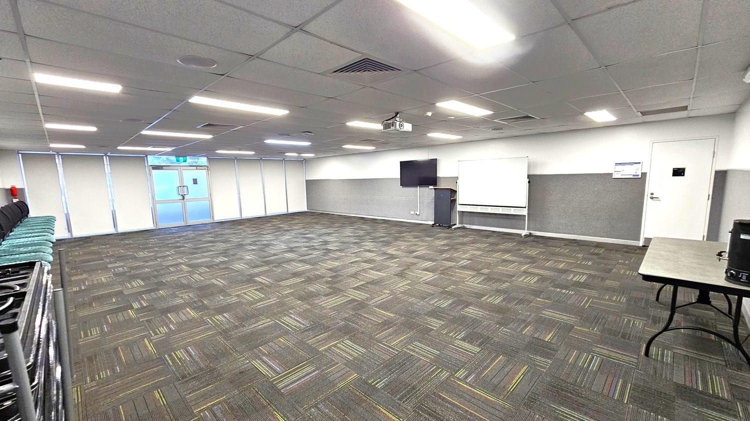 A spacious room with a patterned carpet in shades of grey and green. On the left, there are stacked chairs against the wall. The back wall features large windows with blinds and a glass door. On the right, a whiteboard, a wall-mounted TV, and a lectern are visible. A table with a coffee urn is in the foreground on the right. The ceiling is fitted with fluorescent lights and a projector.