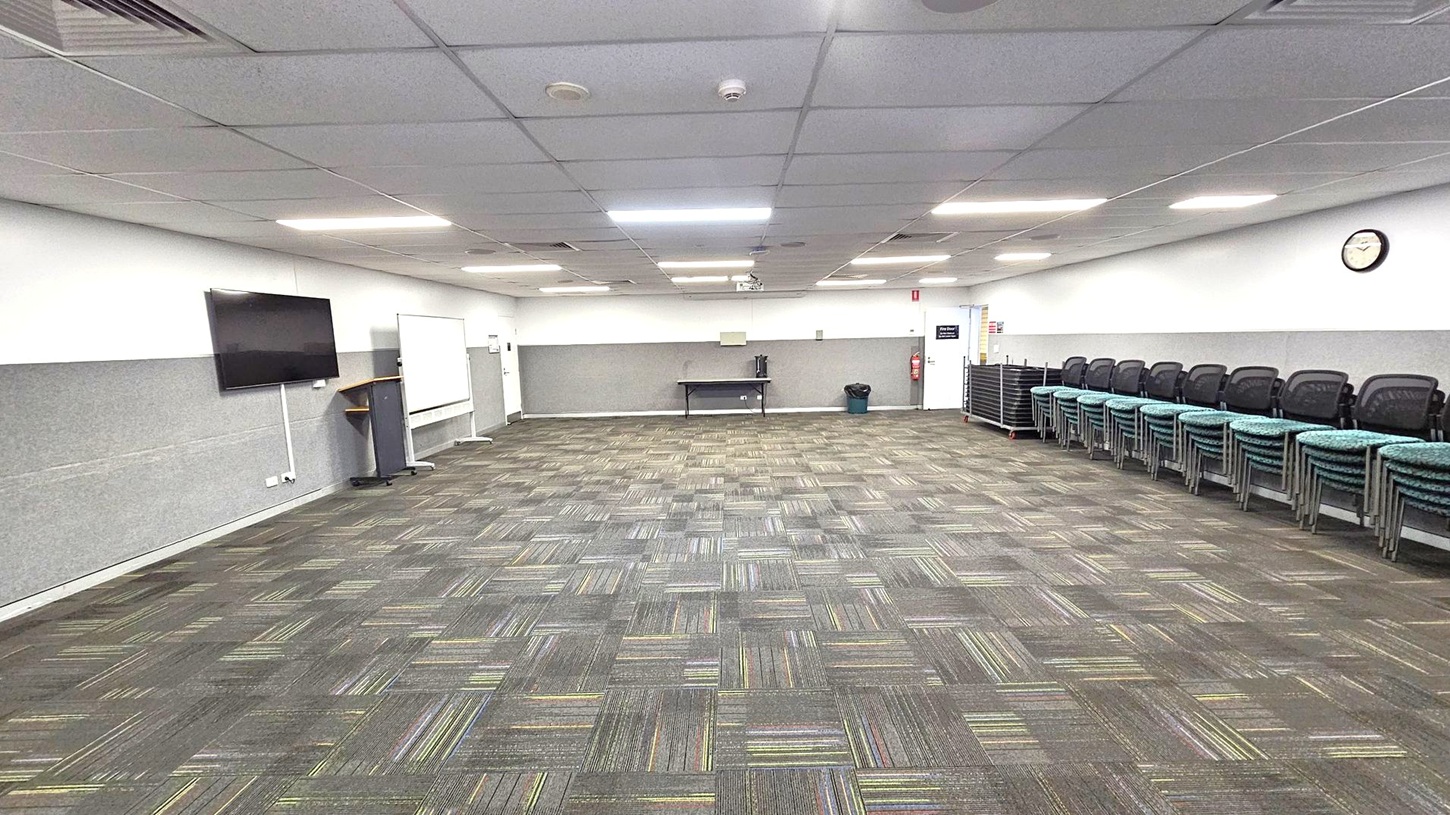 A spacious room with a patterned carpet floor in shades of grey and green. On the left, a wall-mounted TV and a lectern are visible. In the centre, a whiteboard and a table with a kettle are against the wall. On the right, a stack of chairs is neatly arranged, and a clock is mounted on the wall. The ceiling is fitted with fluorescent lights and air vents.