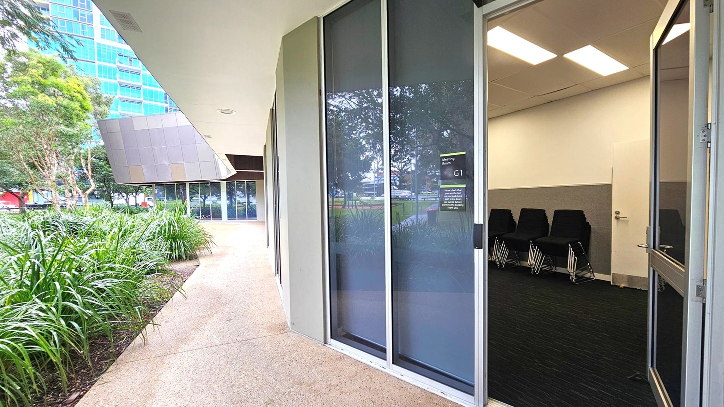 A modern building exterior with a pathway lined by lush green plants on the left. The building has large glass windows and an open door leading to a meeting room labelled "G1" on the right. Inside, several black chairs are stacked against a wall. The ceiling has rectangular fluorescent lights. In the background, a tall building with blue-tinted windows is visible. The floor outside is a textured concrete surface.