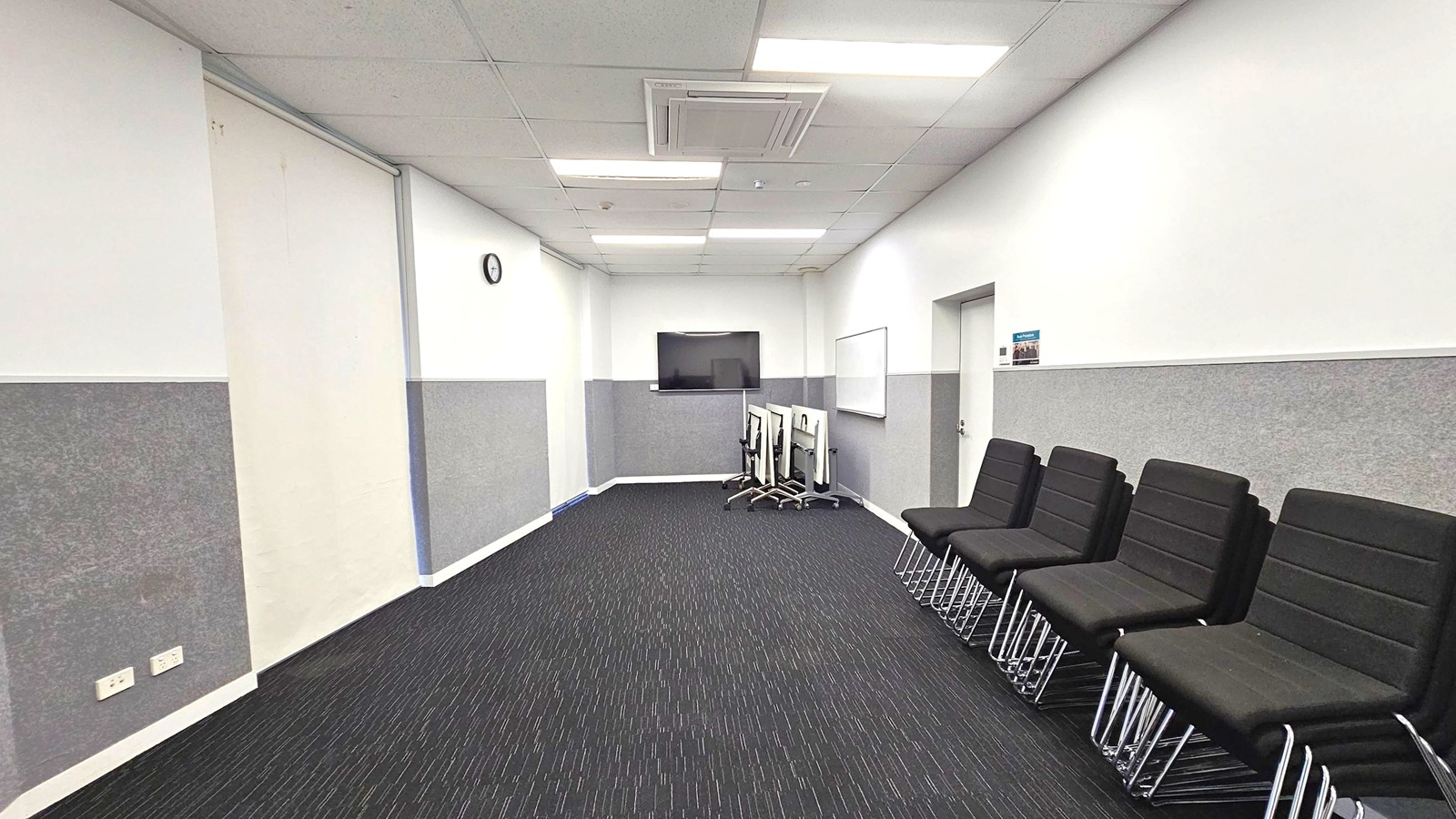 A room with a dark carpeted floor and a white ceiling with fluorescent lights. On the right, there are several black chairs with metal legs lined up against the wall. On the left, there are power outlets on the wall. At the far end, a large flat-screen TV is mounted on the wall, with a whiteboard nearby. A few stacked chairs with wheels are positioned beneath the TV. A clock is mounted on the left wall, and a door is visible on the right wall.