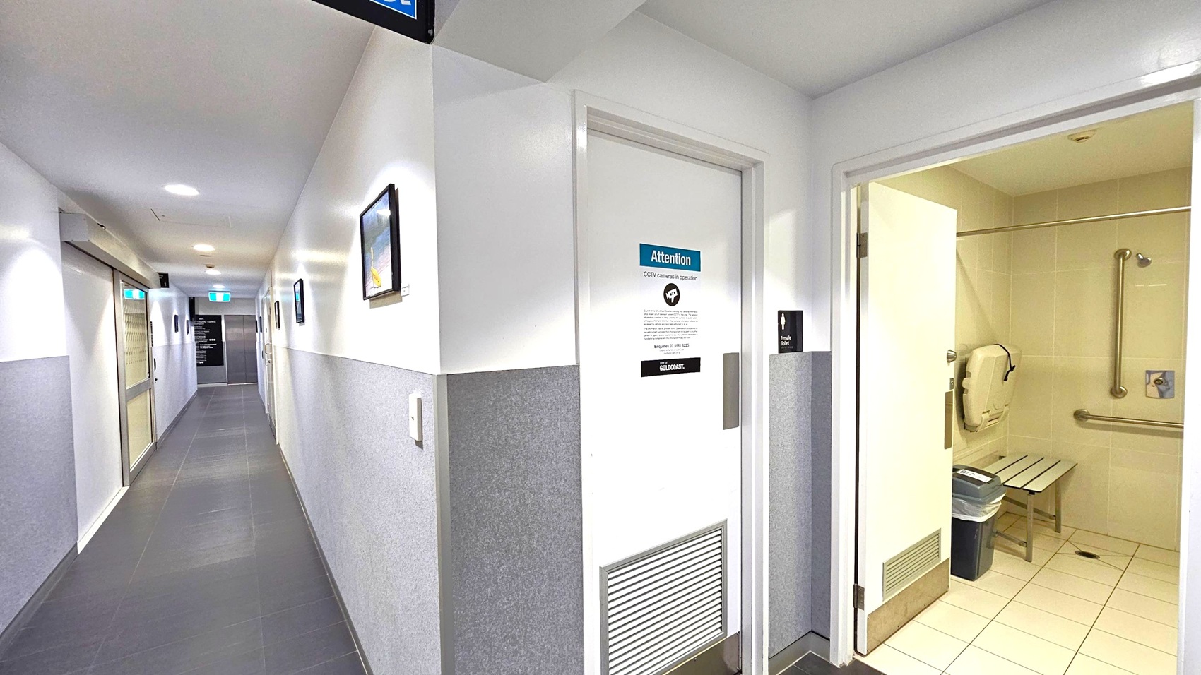A corridor with grey and white walls and a dark tiled floor. On the right, an open door reveals a bathroom with beige tiles, a bench, and grab bars. The door has a sign about CCTV operation. On the left, the corridor extends with framed pictures on the wall and a glass door leading to another area. Ceiling lights illuminate the space, and an exit sign is visible in the distance.