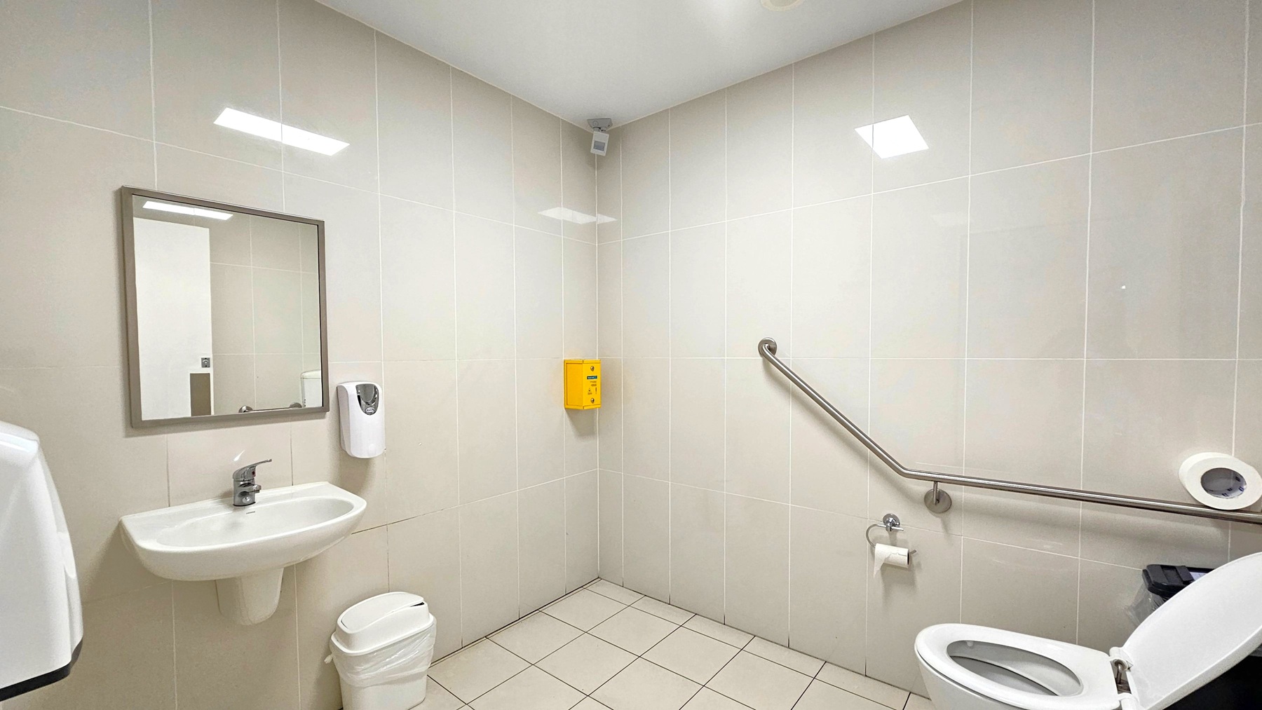 A clean, accessible bathroom with white tiled walls and a light-coloured tiled floor. On the left, there's a wall-mounted sink with a mirror above it and a soap dispenser to the right. Below the sink is a small waste bin. On the right, a toilet with an open lid is equipped with a grab rail for accessibility. A toilet paper holder is mounted on the wall. A yellow box is fixed in the corner above the grab rail. The ceiling is plain white with recessed lighting.