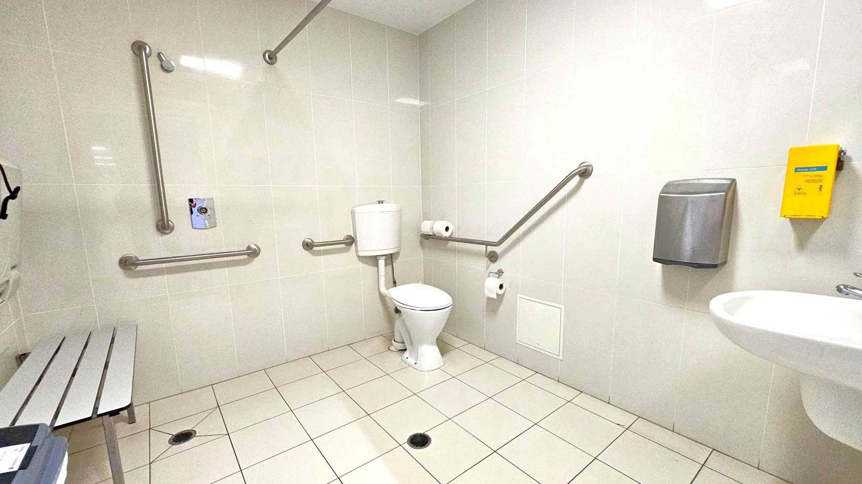 A spacious, accessible bathroom with light-coloured tiled walls and floor. In the centre, a toilet is equipped with multiple metal grab bars on the walls for support. To the right, a wall-mounted sink is visible, along with a stainless steel hand dryer and a yellow emergency call button. On the left, a bench is partially visible. The ceiling is plain and white, with a light fixture.