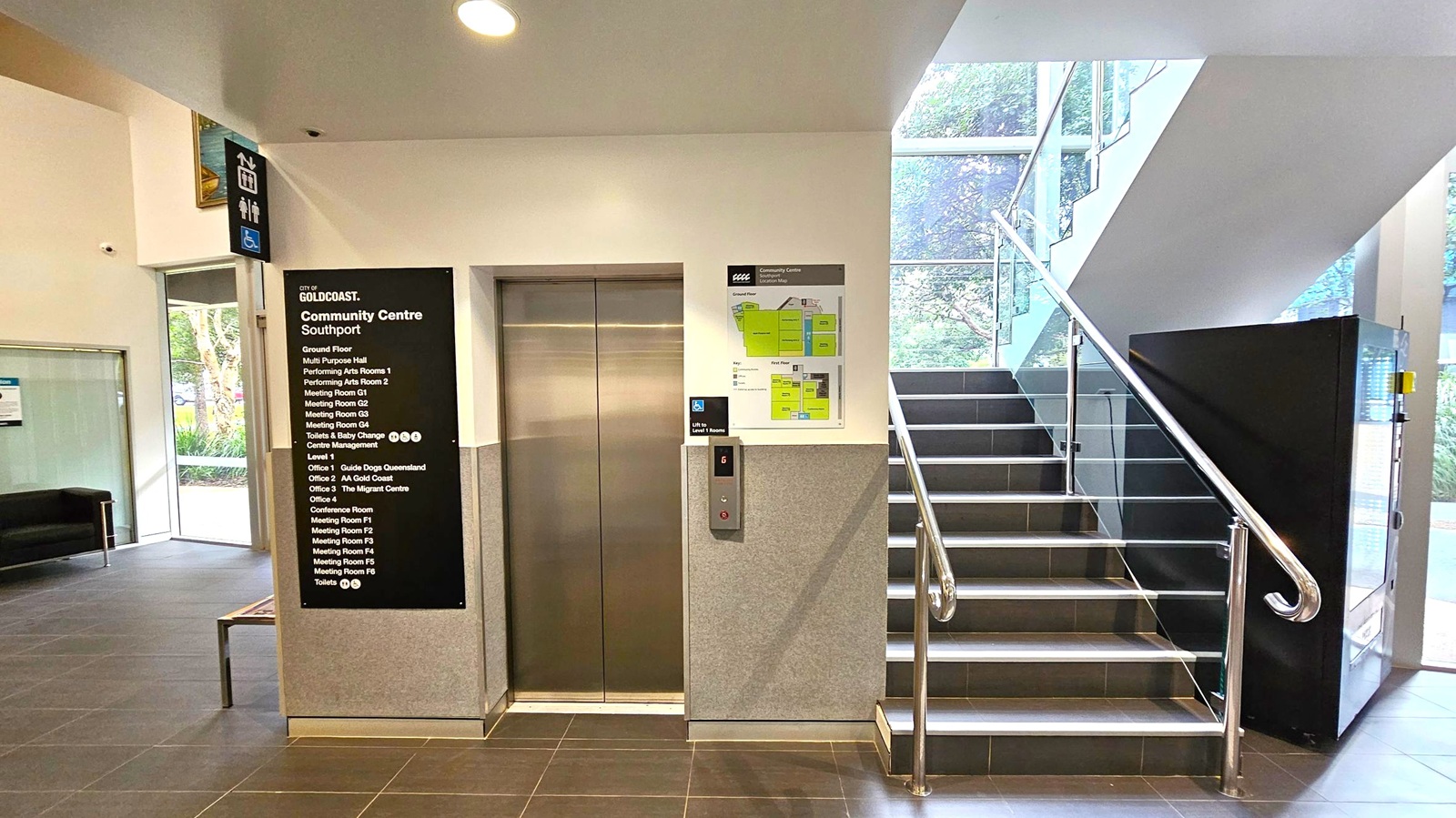 Elevator in the centre with closed metallic doors, flanked by a directory sign listing various rooms and facilities on the left. A staircase with metal railings is on the right, leading upwards. A vending machine is positioned at the base of the stairs. The floor is tiled in dark grey, and the ceiling is white with recessed lighting. Large windows on the right allow natural light to illuminate the area, with greenery visible outside.