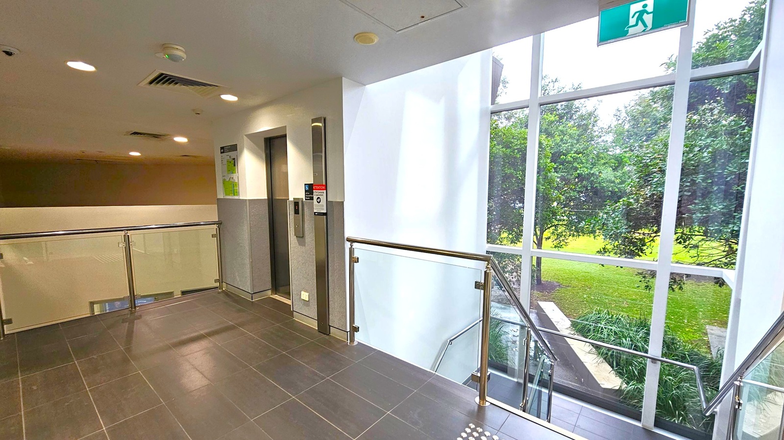 A hallway with dark tiled flooring and a white ceiling featuring recessed lights and vents. On the left, there's a lift with a control panel and a sign above. A glass and metal railing lines the corridor. On the right, large windows reveal a view of green trees and grass outside. An emergency exit sign is visible above the windows.