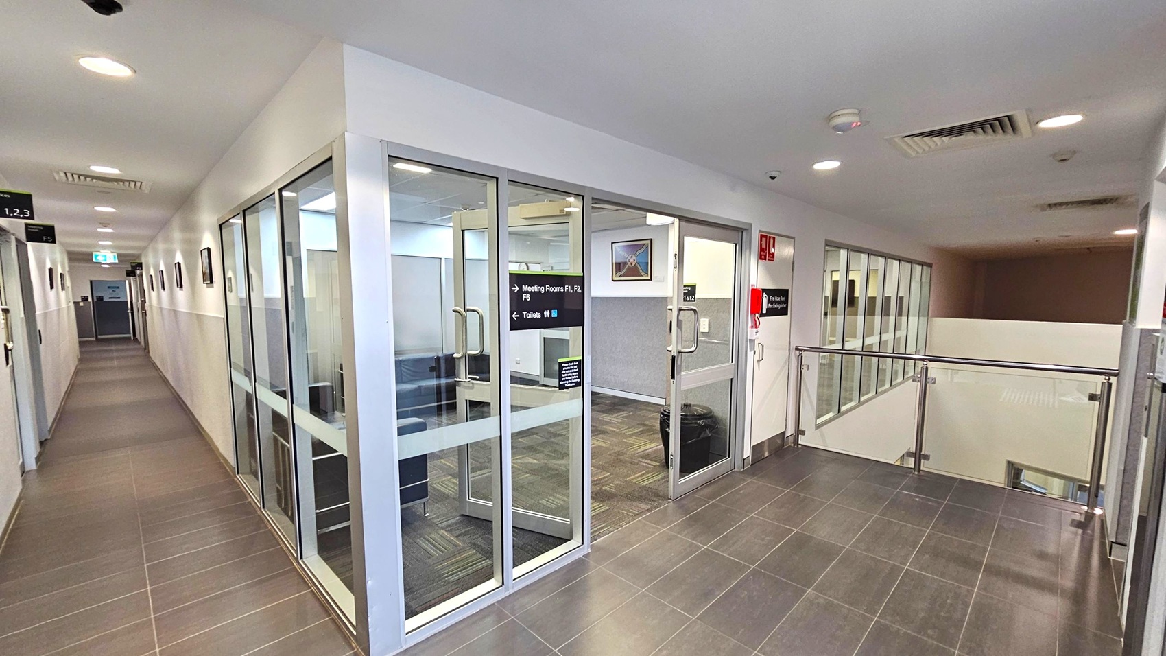 A corridor with a tiled floor and white walls. On the left, a hallway extends with several doors and framed pictures on the wall. On the right, a glass-walled meeting room is visible, with a sign indicating directions to meeting rooms and toilets. The ceiling has recessed lighting and air vents. A railing is present on the right, overlooking a lower level.