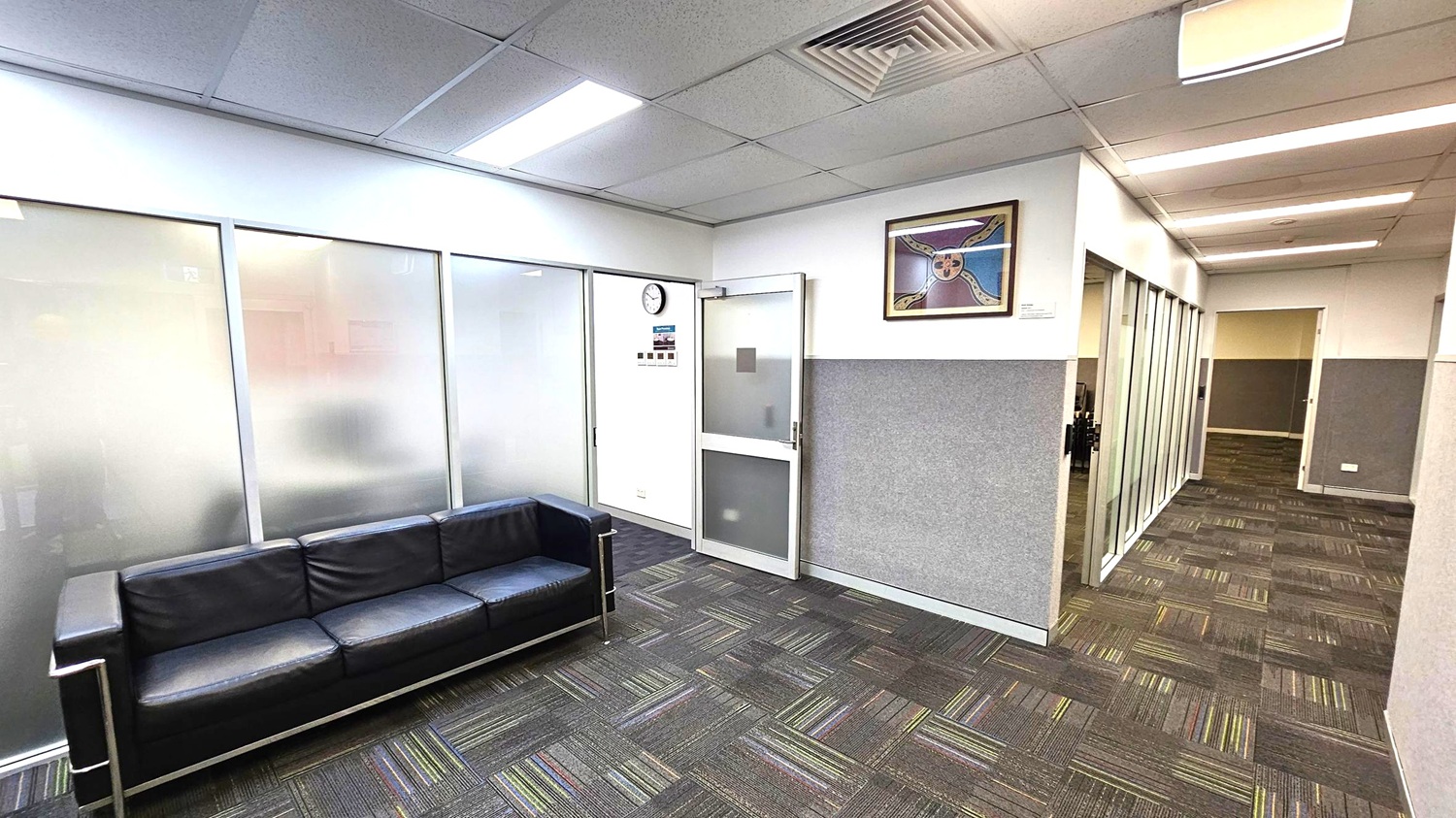 A modern office interior with a black leather sofa on the left against frosted glass panels. The floor is covered with patterned carpet tiles in shades of grey and green. On the right, a corridor with glass partitions leads to other rooms. A wall clock and a colourful framed artwork are mounted on the wall above a door. The ceiling features recessed lighting and air vents.