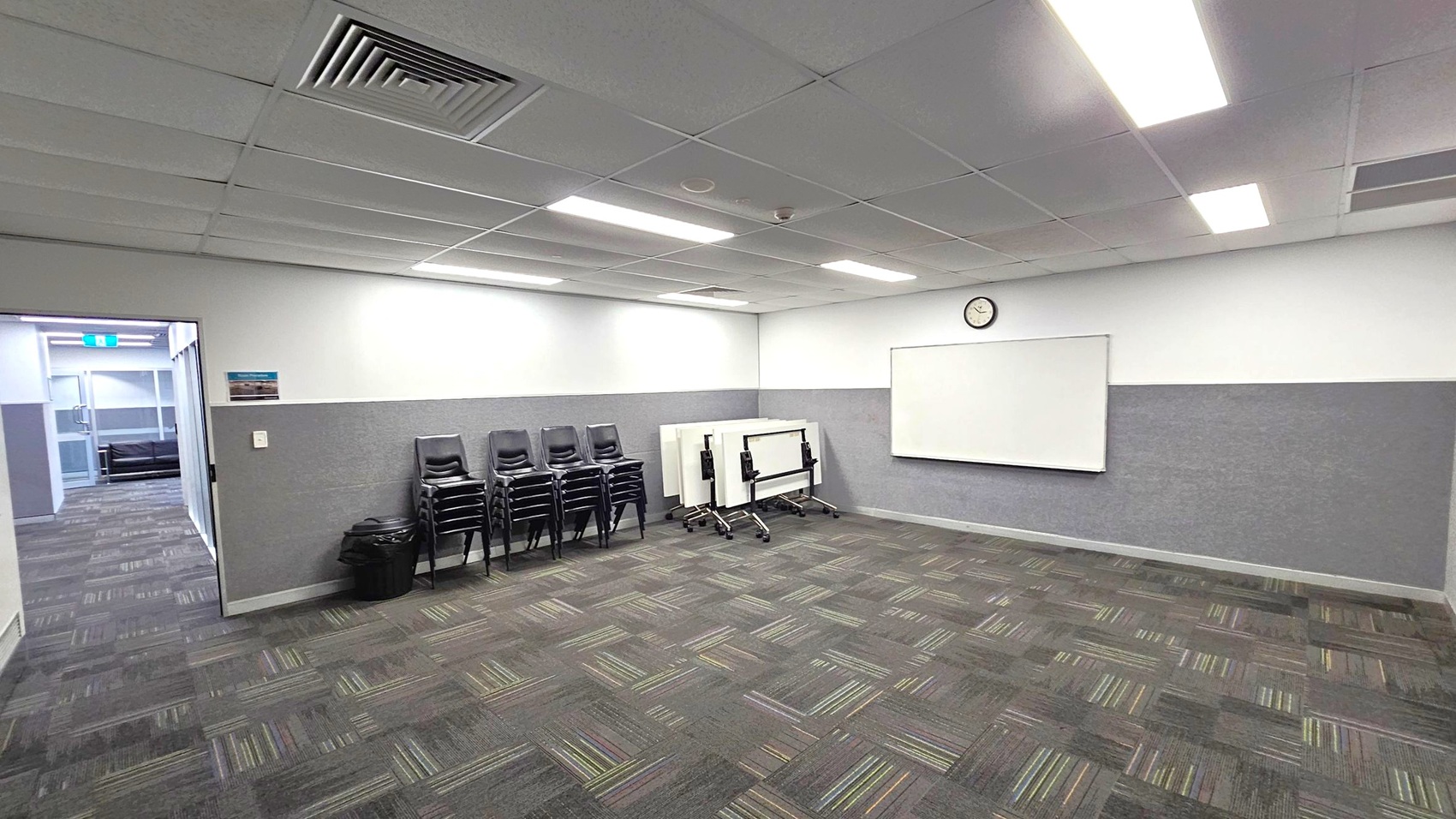 A room with a patterned carpet floor and a tiled ceiling with fluorescent lights. On the right wall, there's a whiteboard and a clock. In the middle, several stacked chairs and folded tables are against the wall. On the left, an open doorway leads to a corridor with more patterned flooring and a visible exit sign. A small bin is near the chairs.