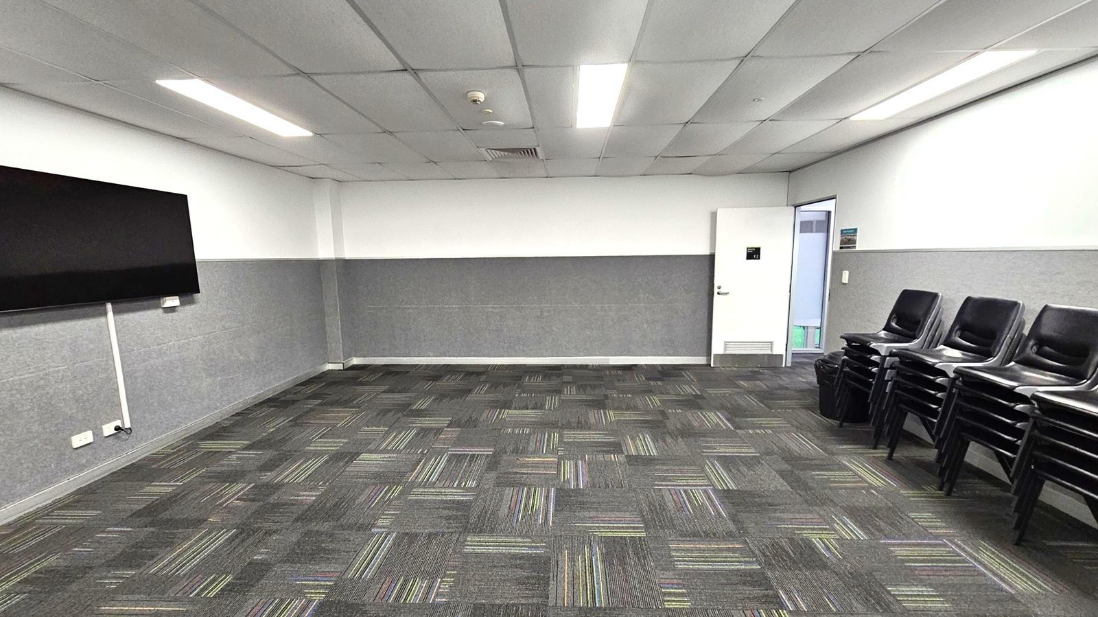 A room with a patterned carpet floor featuring grey and green stripes. On the left, a large flat-screen TV is mounted on the wall. On the right, there are stacks of black chairs. The back wall has a door slightly ajar, revealing a glimpse of another room. The ceiling is fitted with rectangular fluorescent lights. The walls are half-panelled with grey and white.