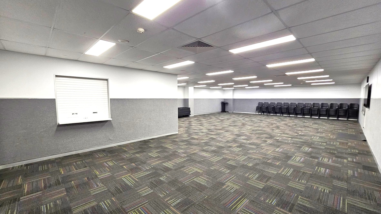 A large, empty room with a patterned carpet featuring grey and green tones. The ceiling is lined with rectangular fluorescent lights. On the left wall, there's a closed shutter window. The right side has a stack of black chairs lined up against the wall. In the background, more chairs are stacked, and a bin is visible. The walls are two-toned, with a darker shade on the lower half.