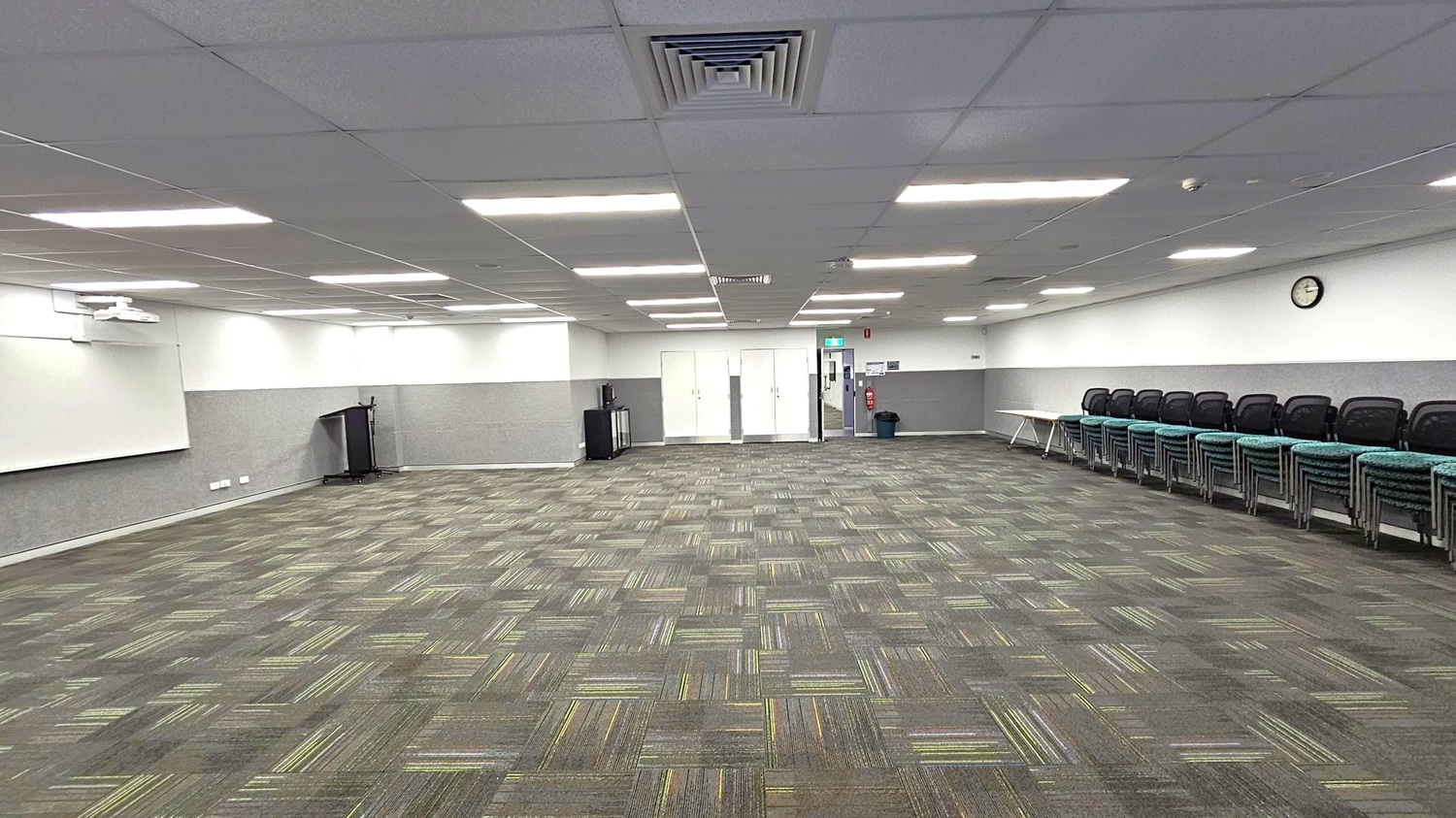 A spacious room with a patterned carpet in shades of green and grey. On the left, there's a whiteboard and a podium. The back wall features double doors and a small cabinet. On the right, a row of stacked chairs lines the wall beneath a clock. The ceiling is fitted with fluorescent lights and ventilation panels.