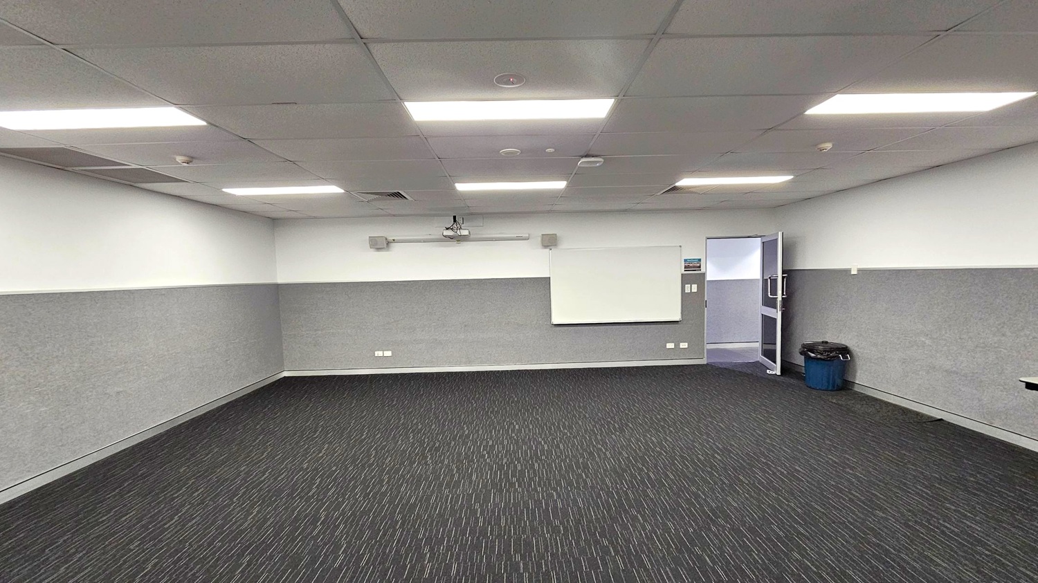 A room with a dark carpeted floor featuring a subtle linear pattern. The walls are light grey with a whiteboard mounted on the centre of the far wall. Above the whiteboard, a projector is installed. On the right, an open door leads to another area, and a blue bin is placed nearby. The ceiling is fitted with rectangular fluorescent lights, evenly spaced across the room.