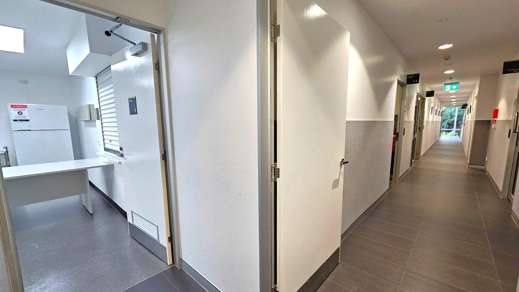 A corridor with a tiled floor and white walls. On the left, an open door reveals a small room with a white counter, a refrigerator, and a sign on the wall. The corridor extends to the right, lined with closed doors and lit by ceiling lights. An exit sign is visible above a door at the end of the hallway, leading to a glass door with a view of greenery outside.