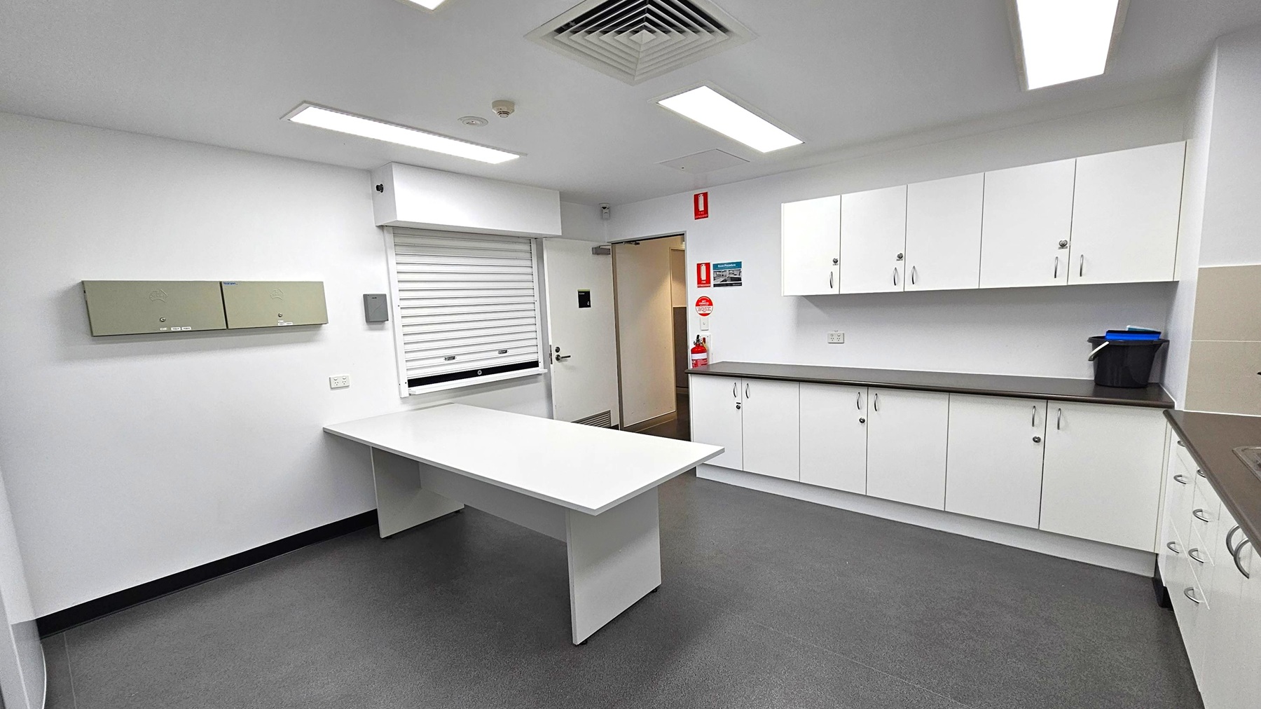 A clean, modern room with white walls and grey flooring. On the left, there are two light green wall-mounted cabinets. In the centre, a white table extends from the wall. The right side features a long countertop with white cabinets above and below. A small black bin and some blue items are on the counter. A door with a fire extinguisher beside it is in the background. The ceiling has recessed lighting and a ventilation grille.