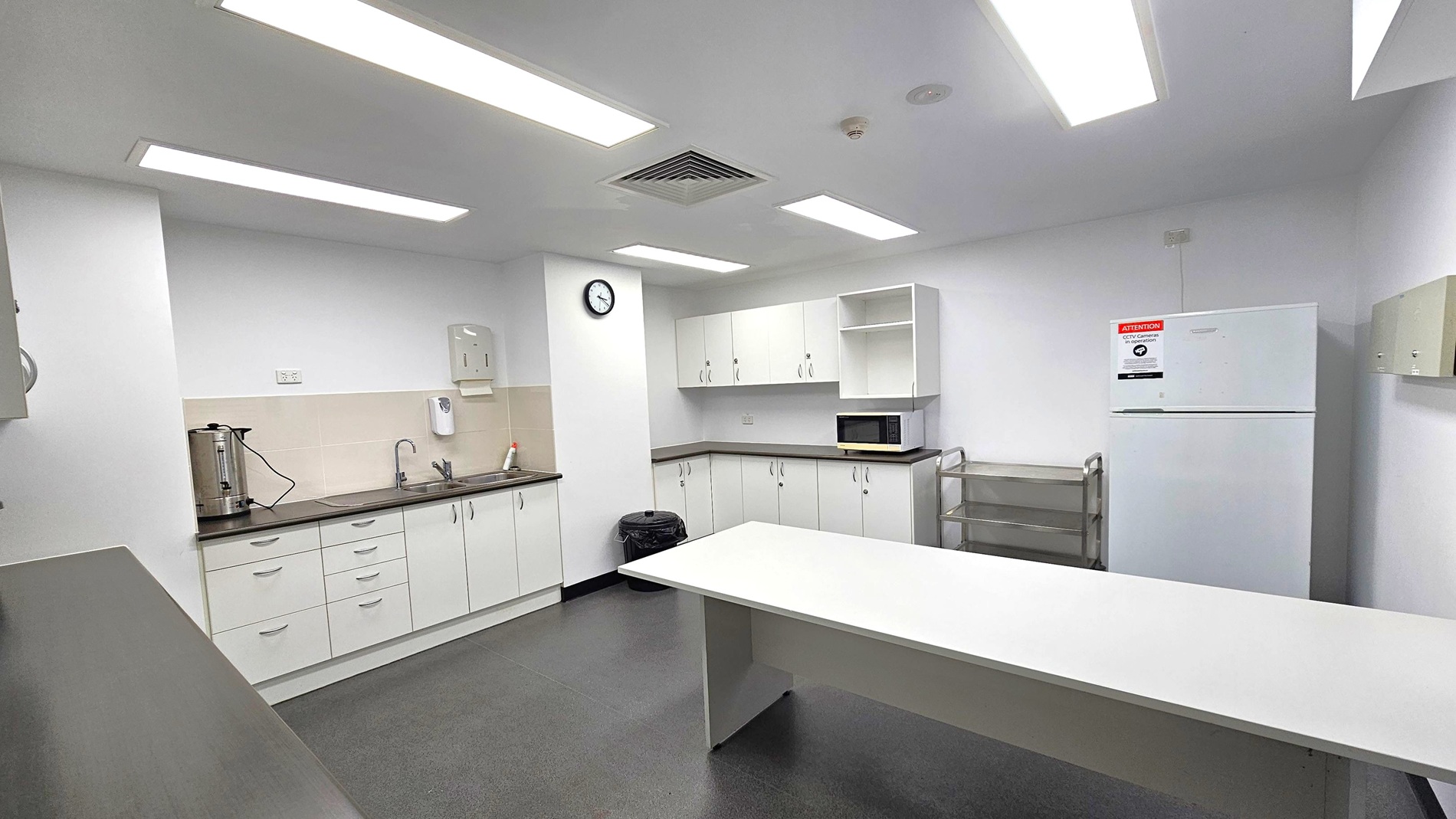 A clean, modern kitchen with white cabinetry and a dark countertop. On the left, a sink with a kettle and soap dispenser is visible. A wall clock is mounted above. In the centre, a white table stands on a dark floor. On the right, a microwave sits on a counter, with a refrigerator nearby. The ceiling has several rectangular fluorescent lights and a vent. A small metal trolley is positioned next to the fridge.