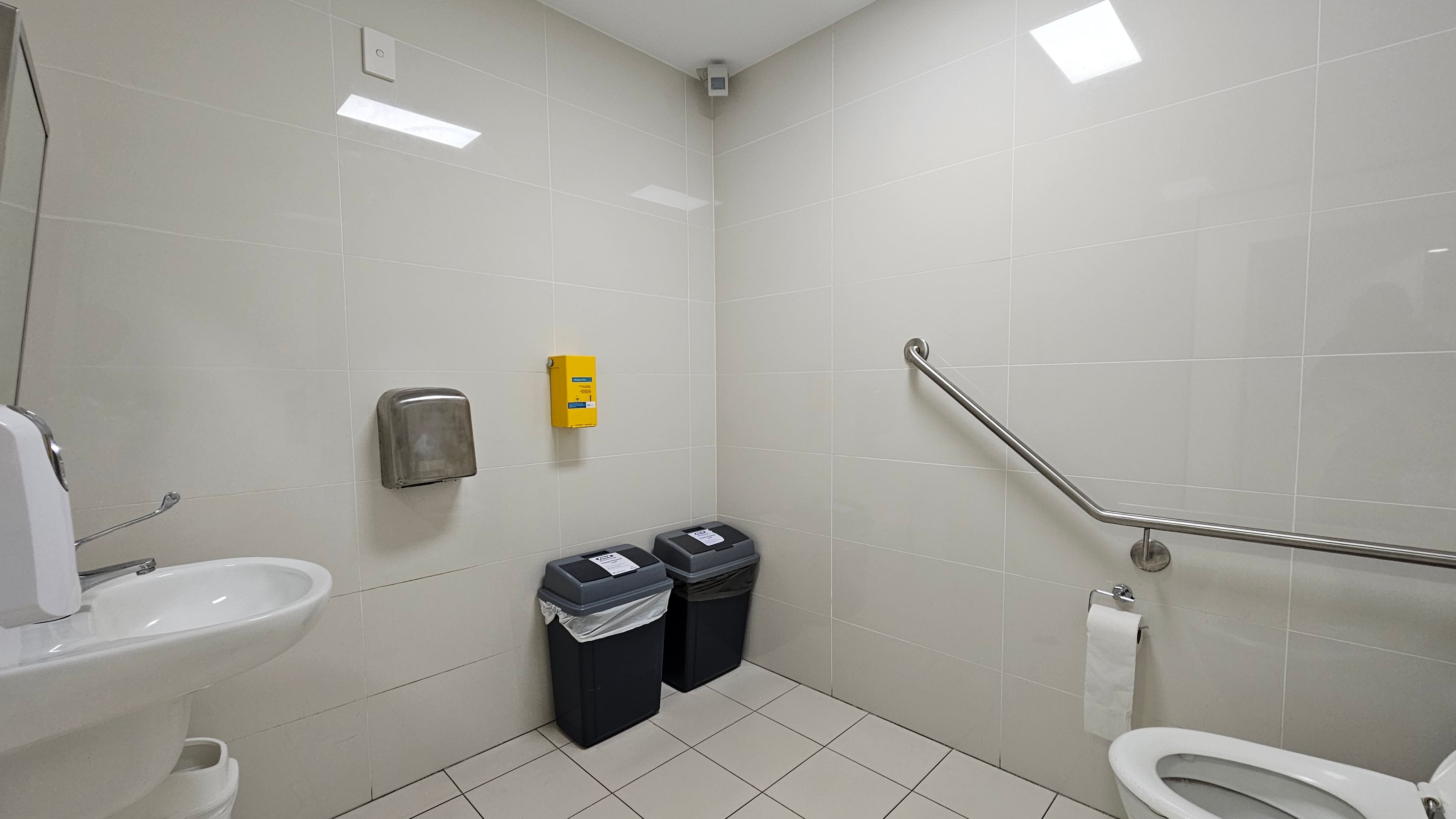A public restroom with white tiled walls and floor. On the left, a white sink with a soap dispenser and a hand dryer above it. In the centre, two black bins with lids are placed side by side beneath a yellow wall-mounted box. On the right, a toilet with a roll of toilet paper on a holder and a metal grab bar above it. The ceiling is plain with a light fixture.