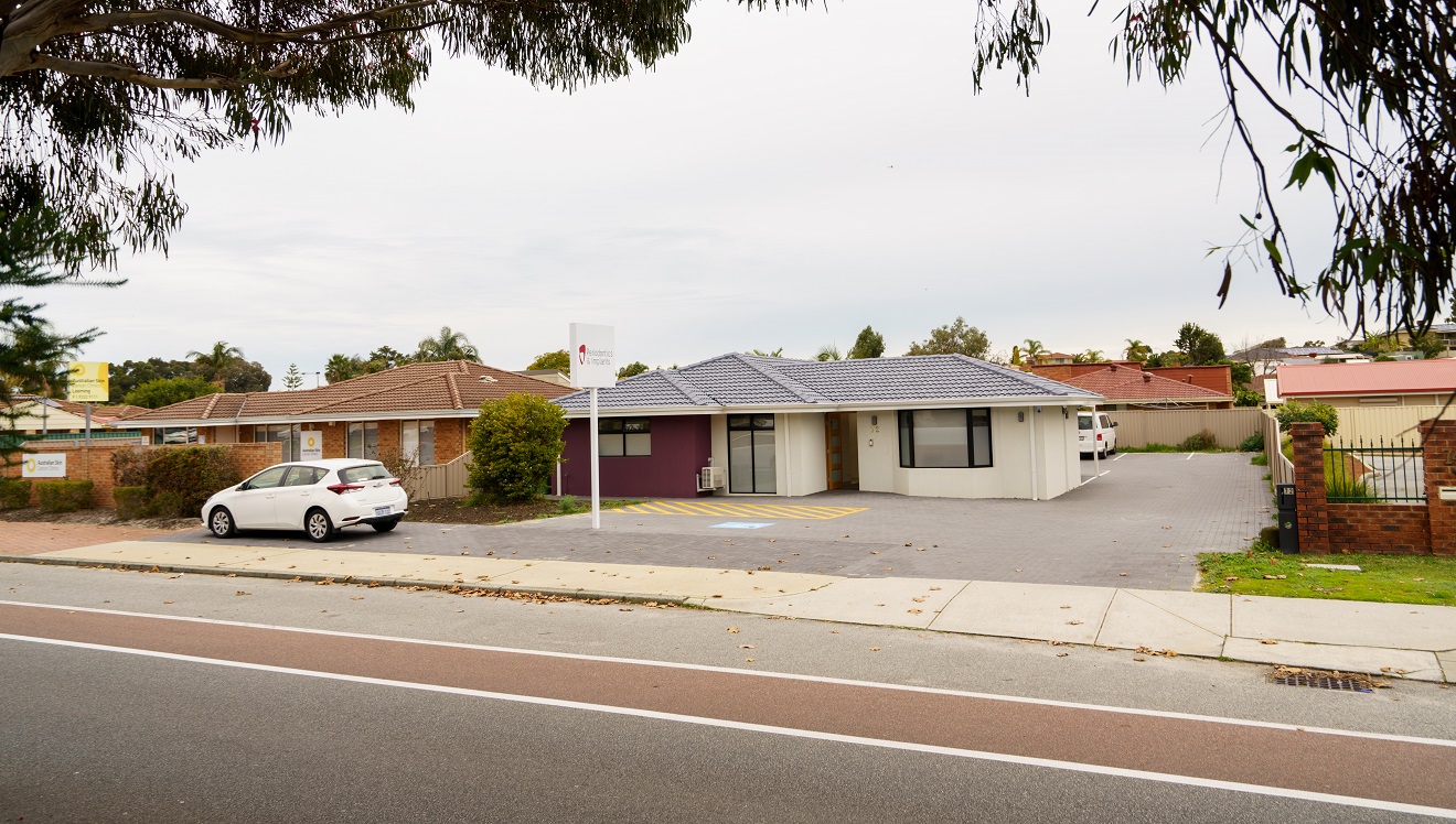 A cream building with a grey, tiled roof with a feature wall to the left in purple. The front door is timber with four glass panels. To the left is a white sign with 'Periodontics & Implants'. To the right is a glimpse of a small, white van parked behind the building and a sall white vehicle is parked out the front of the building. An accessible car parking space is directly adjacent to the front entrance. Other suburban style houses and businesses surround the business. Trees frame the view. A cream building with a grey, tiled roof with a feature wall to the left in purple. The front door is timber with four glass panels. To the left is a white sign with 'Periodontics & Implants'. To the right is a glimpse of a small, white van parked behind the building and a sall white vehicle is parked out the front of the building. An accessible car parking space is directly adjacent to the front entrance. Other suburban style houses and businesses surround the business. Trees frame the view.