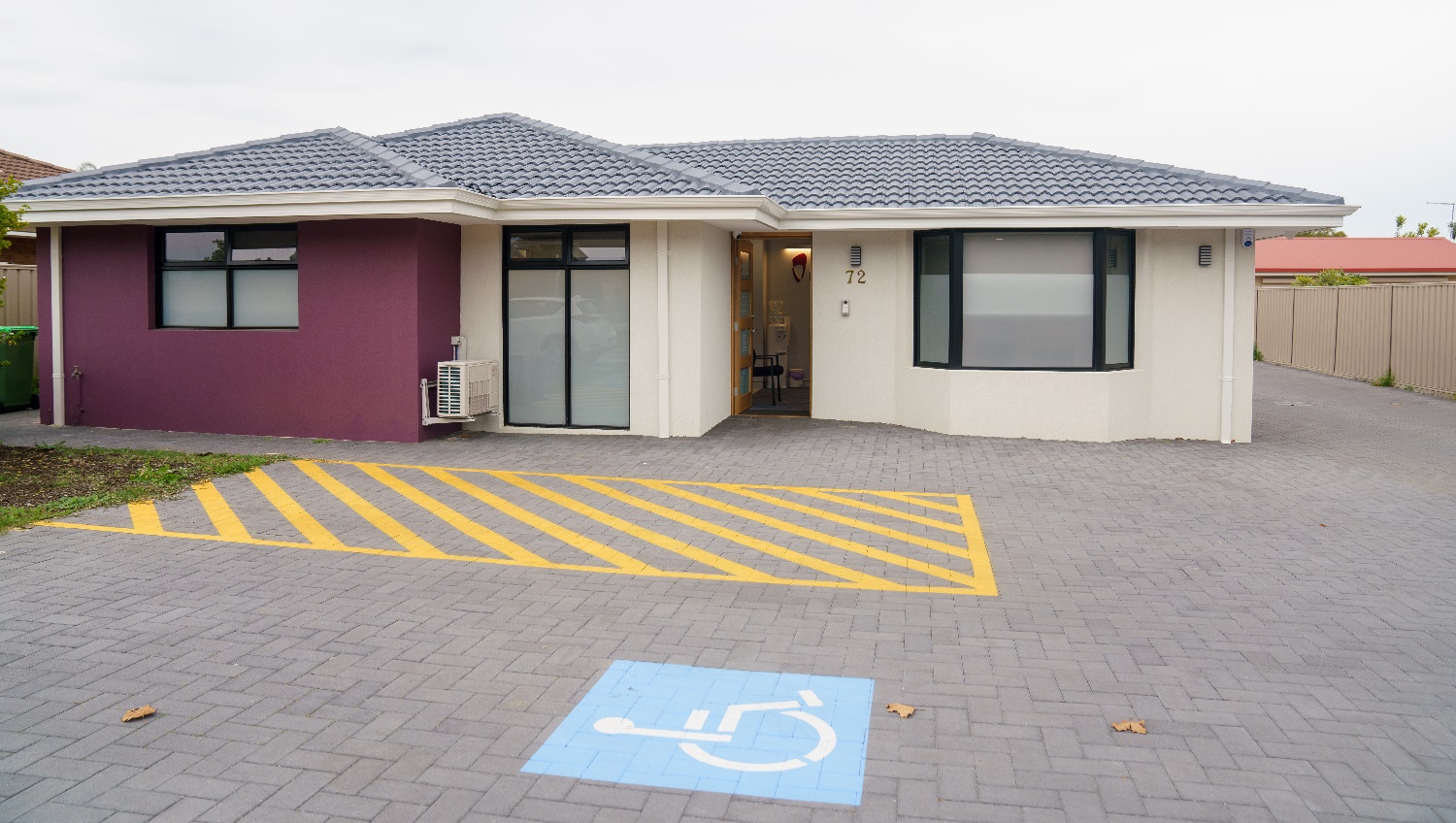 A closer look at the cream building with a grey, tiled roof with a feature wall to the left in purple. The front door is timber with four glass panels and is open with a glimpse of a chair is visible inside. A cream colourbond fence sits t the right and a red roof is visible to the right and behind this business. An accessible car parking space is directly adjacent to the front entrance. A closer look at the cream building with a grey, tiled roof with a feature wall to the left in purple. The front door is timber with four glass panels and is open with a glimpse of a chair is visible inside. A cream colourbond fence sits t the right and a red roof is visible to the right and behind this business. An accessible car parking space is directly adjacent to the front entrance.