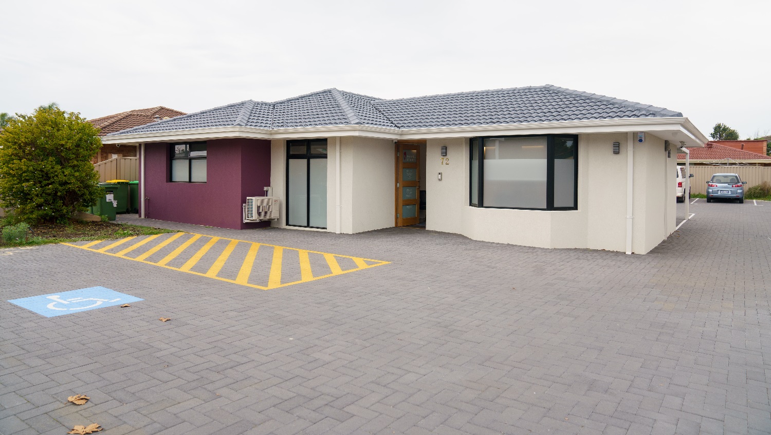 Another angle of a view of the cream building with a grey, tiled roof with a feature wall to the left in purple. The front door is timber with four glass panels. To the left is a white sign with 'Periodontics & Implants'. To the right is a glimpse of a small, white van and a small grey vehicle parked behind the building. An accessible car parking space is directly adjacent to the front entrance. To the left of this buiding are the rubbish bins and a glimpse of the building located next door. Another angle of a view of the cream building with a grey, tiled roof with a feature wall to the left in purple. The front door is timber with four glass panels. To the left is a white sign with 'Periodontics & Implants'. To the right is a glimpse of a small, white van and a small grey vehicle parked behind the building. An accessible car parking space is directly adjacent to the front entrance. To the left of this buiding are the rubbish bins and a glimpse of the building located next door.