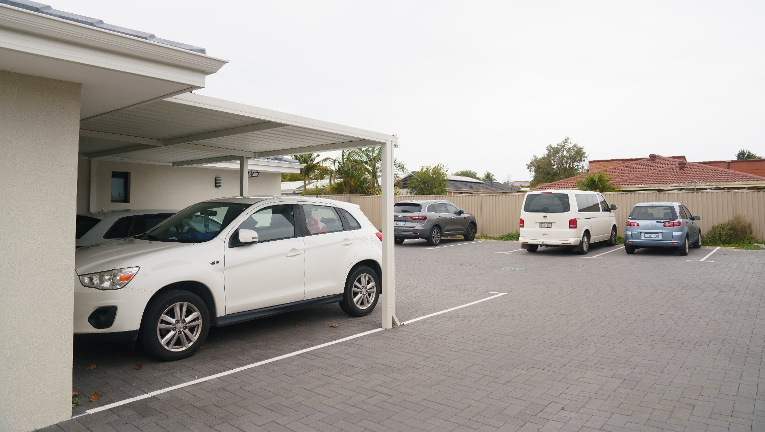 To the left are two white vehicles parked underneath a carport at the back of the cream building. Adjacent are is a small grey vehicle, a white van and a small silver hatchack parked up against the back fence. A glimpse of the house behind is visible and features a red tiled roof. Trees are including palm trees are visible in the backyard of the house located at the back of the business. To the left are two white vehicles parked underneath a carport at the back of the cream building. Adjacent are is a small grey vehicle, a white van and a small silver hatchack parked up against the back fence. A glimpse of the house behind is visible and features a red tiled roof. Trees are including palm trees are visible in the backyard of the house located at the back of the business.
