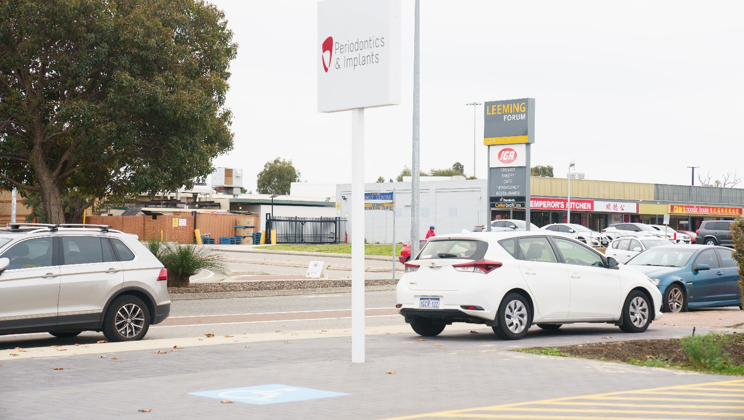 A silver vehicle is parked to the left on the roadside adjacent to a tree, whilst to the right a small white hatchback is parked outside the business underneath a white sign stating 'Periodontics & Implants'. Across the road a number of business are located at the 'Leeming Forum' including an IGA, Emperoros Kitchen and Noodle House. Vehicles are parked outside this retail precinct. A silver vehicle is parked to the left on the roadside adjacent to a tree, whilst to the right a small white hatchback is parked outside the business underneath a white sign stating 'Periodontics & Implants'. Across the road a number of business are located at the 'Leeming Forum' including an IGA, Emperoros Kitchen and Noodle House. Vehicles are parked outside this retail precinct.