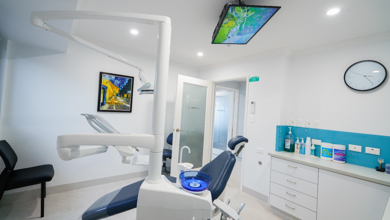 The clinic is neat and organized, with white walls and a sky-blue feature wall; the clinic has four drawers near the door. There is a reclining examination chair that's dark blue in color. This chair appears comfortable and is attached to the basic equipment used by dentists/periodontists such as chairside spittoon, suction, the LED lamp and the like. There is a LED TV mounted in the ceiling above and a painting posted on the wall that used bright and vibrant shades of blue and orange. The clinic is neat and organized, with white walls and a sky-blue feature wall; the clinic has four drawers near the door. There is a reclining examination chair that's dark blue in color. This chair appears comfortable and is attached to the basic equipment used by dentists/periodontists such as chairside spittoon, suction, the LED lamp and the like. There is a LED TV mounted in the ceiling above and a painting posted on the wall that used bright and vibrant shades of blue and orange.